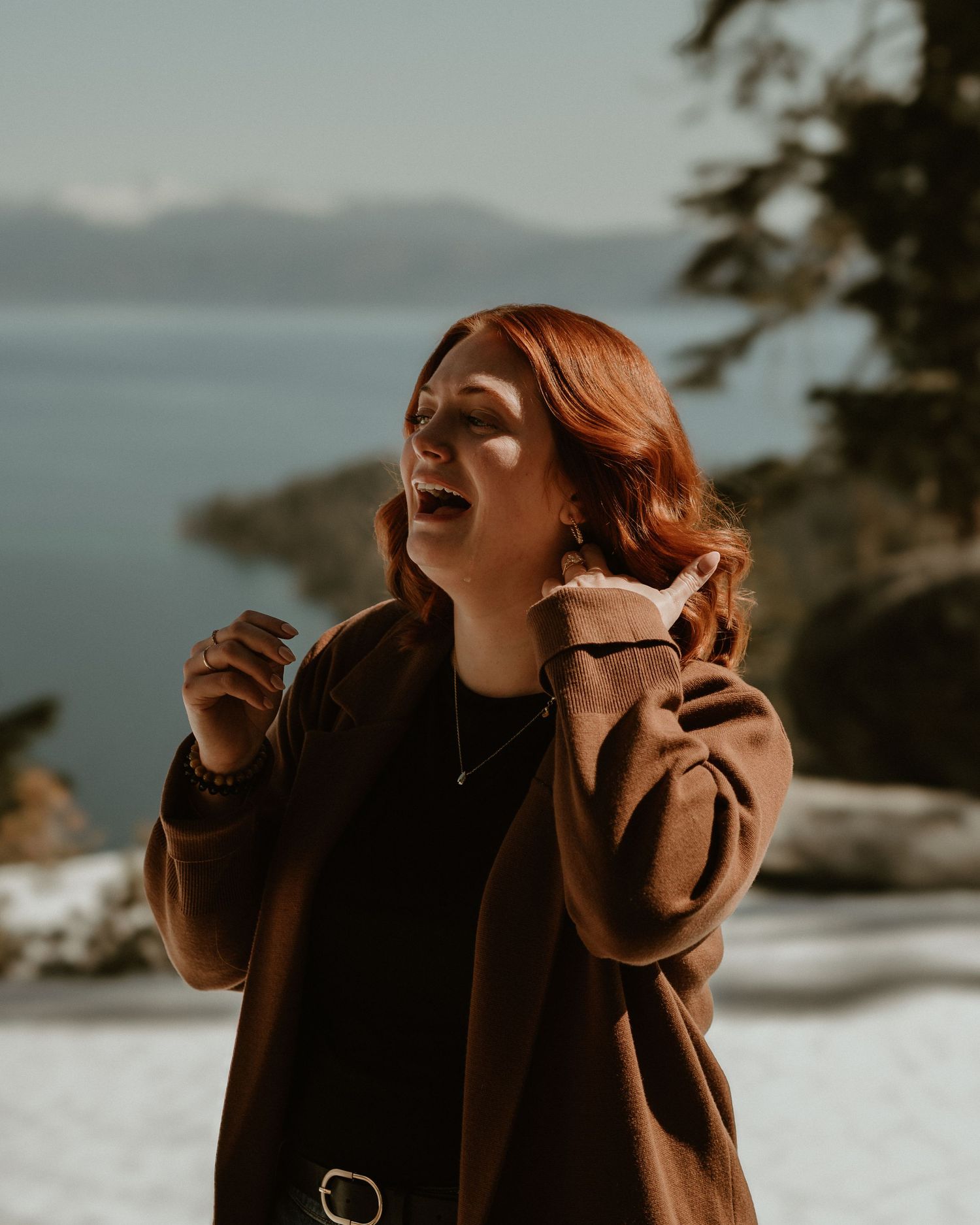 Person in brown coat laughing while holding coffee cup against scenic mountain backdrop.