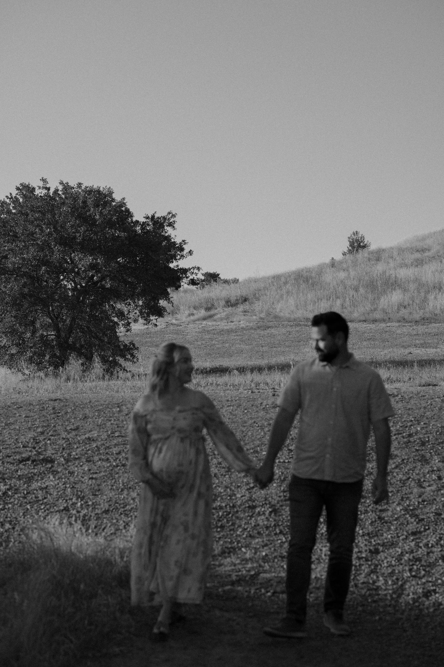 Black and white photo of couple holding hands in a rural field with mountains in background.