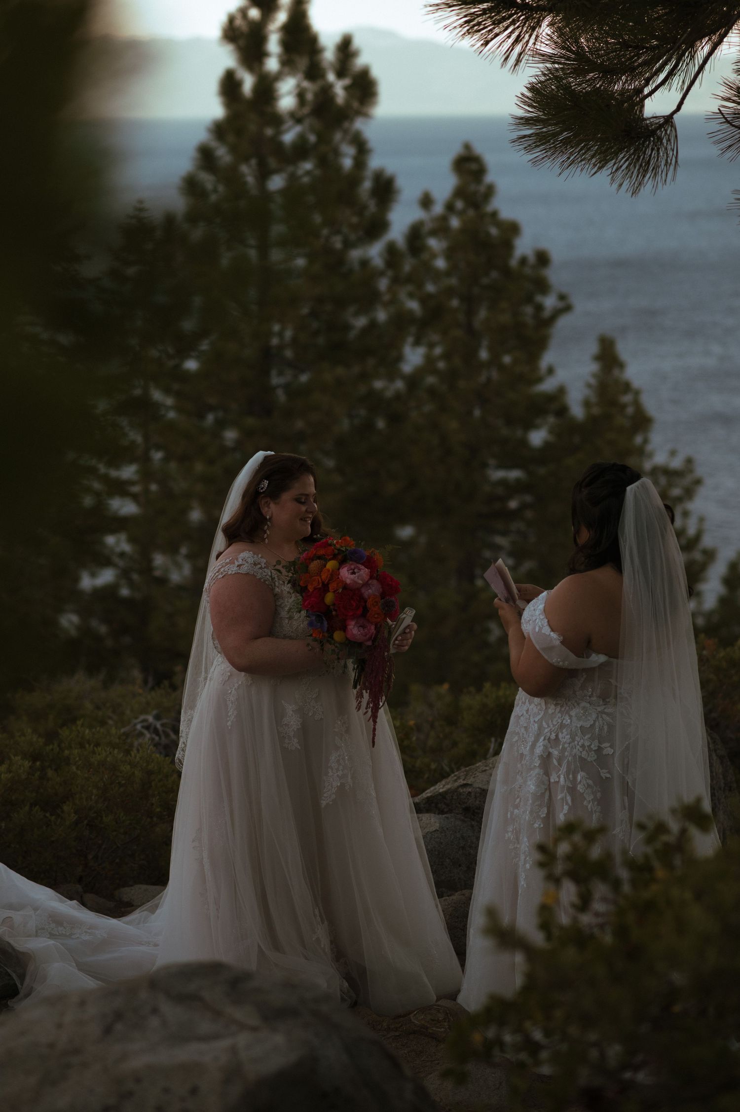 Two brides in white wedding gowns and veils exchange vows outdoors near Lake Tahoe surrounded by pine trees.