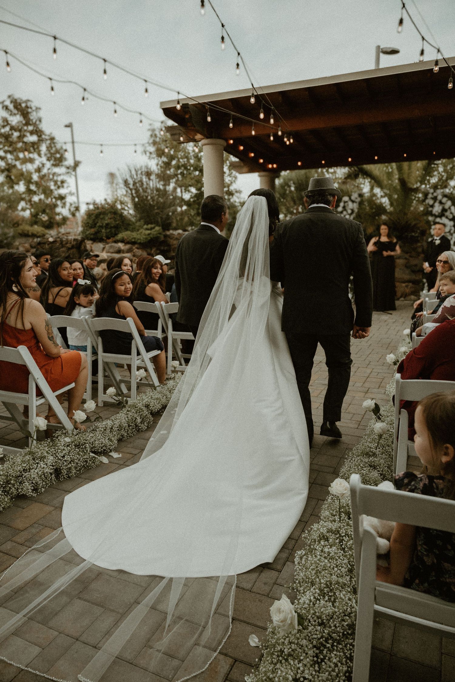 An outdoor wedding ceremony setup with string lights and white chairs lining a flower-decorated aisle.