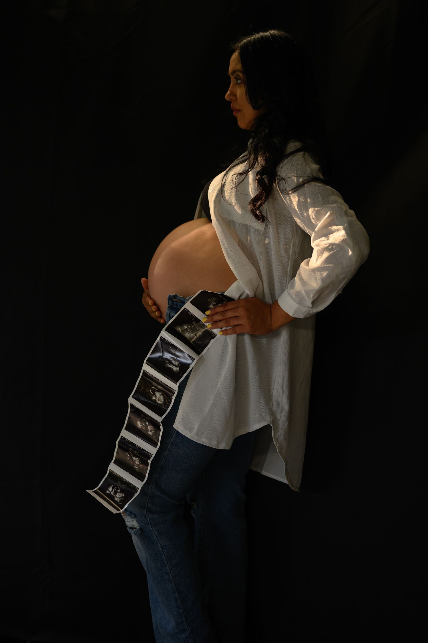Side profile of an expectant mother holding ultrasound photos in a white shirt against dark backdrop.
