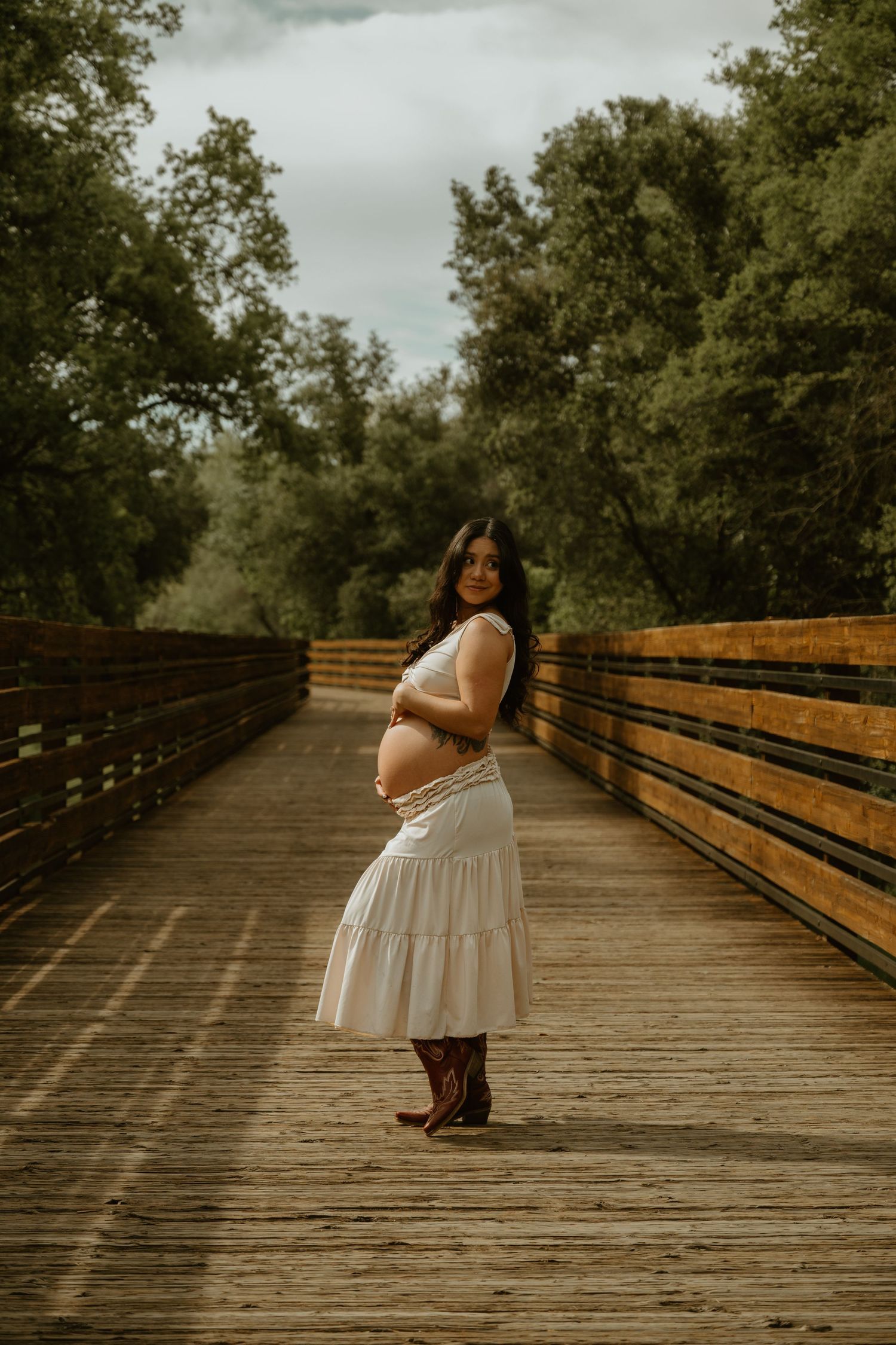 Maternity photo on a wooden bridge with lush green trees in the background.