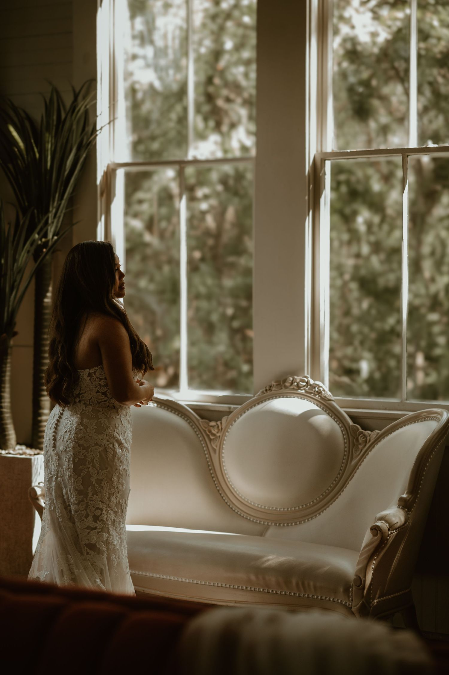 Bride in lace dress standing by vintage white sofa near bright window.