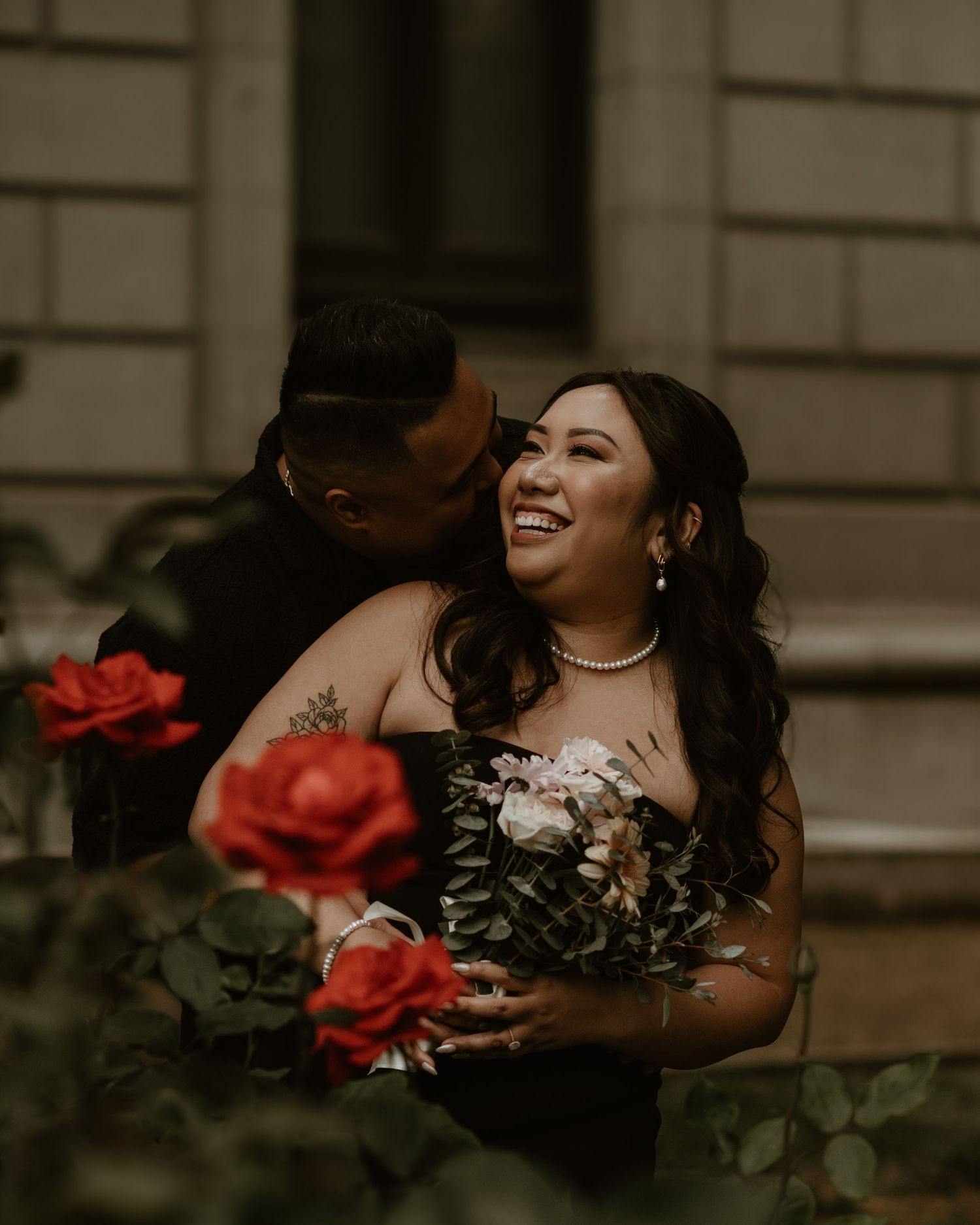 A romantic moment with red roses in foreground as the couple embraces outside a historic building.