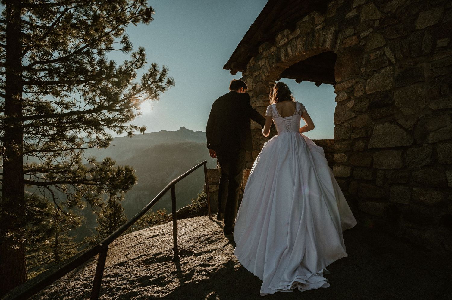 Bride in flowing white dress poses against stone architecture with mountain views.