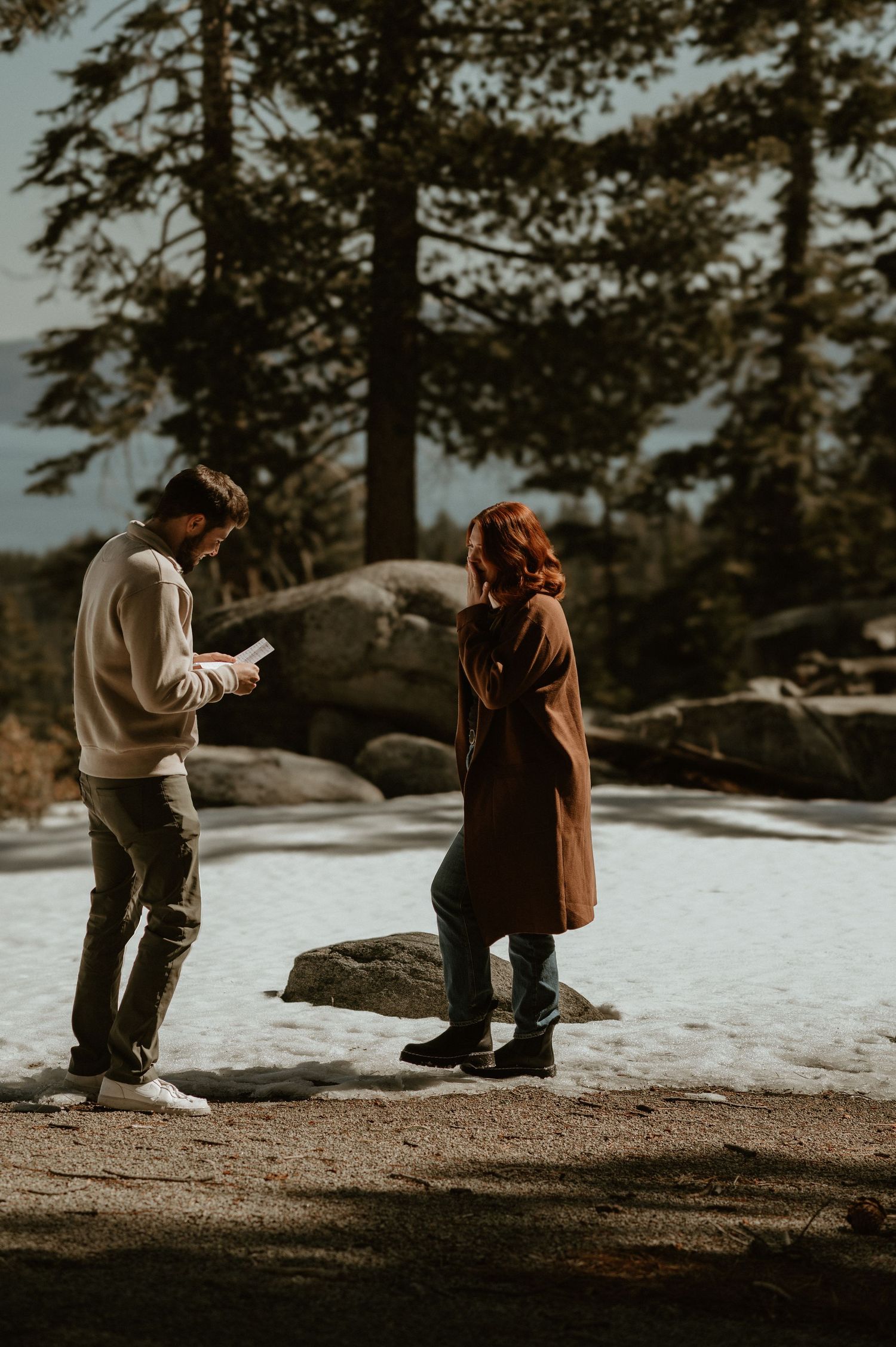 Two people standing in the snow with mountain views and tall evergreen trees.
