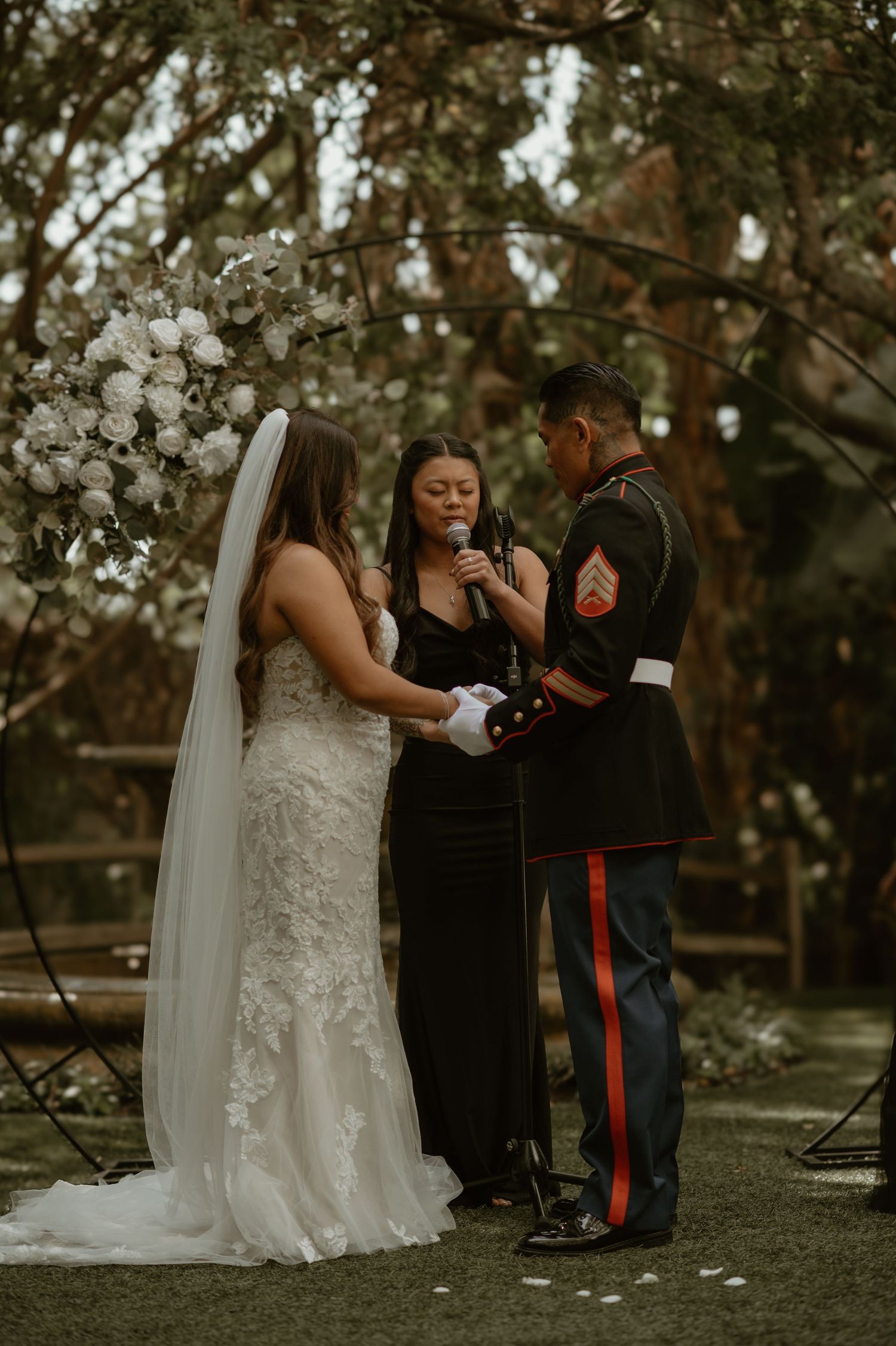 Wedding ceremony in progress under trees with officiant and couple exchanging vows.