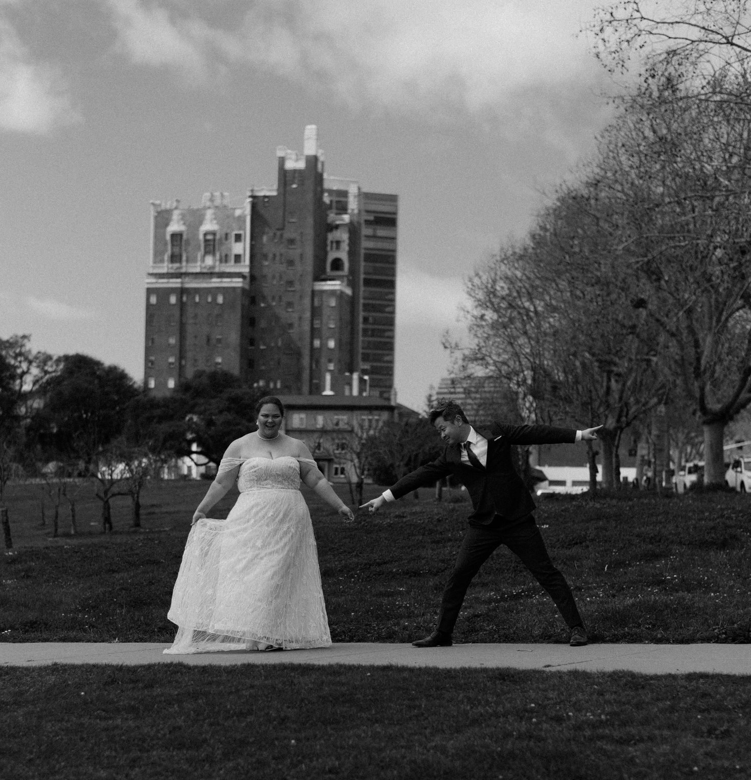 A playful wedding photo with couple striking dramatic poses on grass with historic building behind.