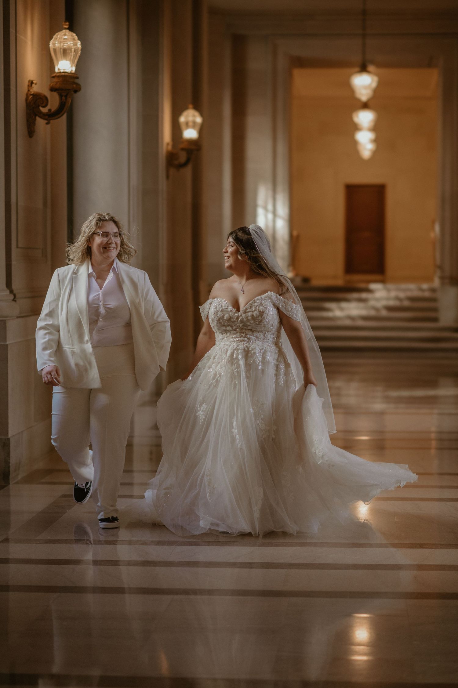 Two brides walk hand in hand through an elegant chandelier-lit hallway, one in a suit and one in a ballgown.