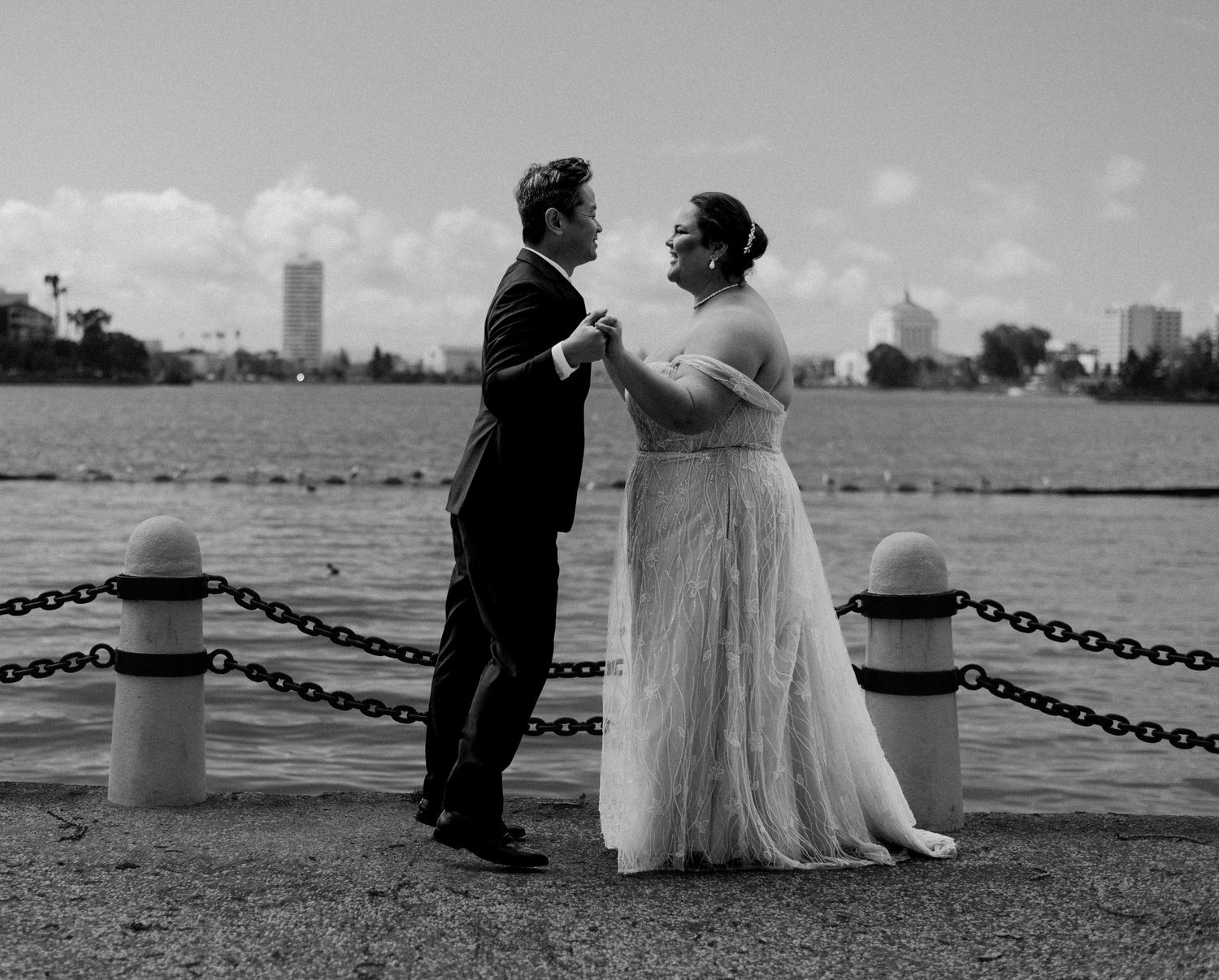 A couple shares a romantic moment by the waterfront with city buildings in the background.
