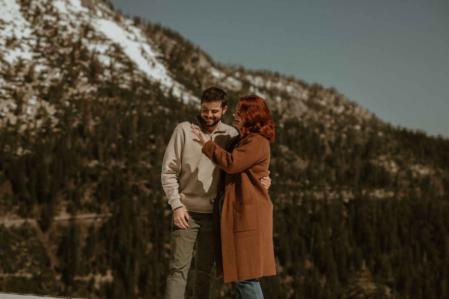 A couple embraces against a backdrop of snow-capped mountains and evergreen forest in moody lighting.