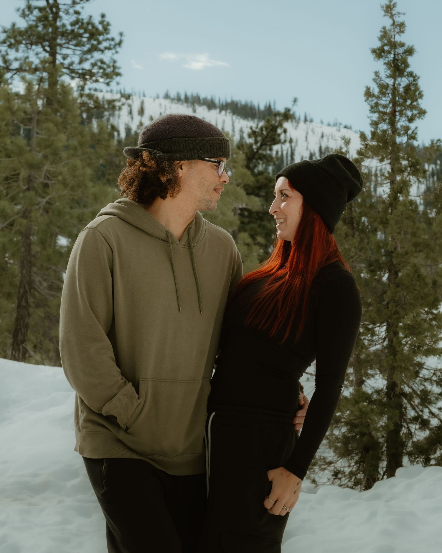 A couple shares a tender moment in a snowy forest landscape wearing casual winter attire.
