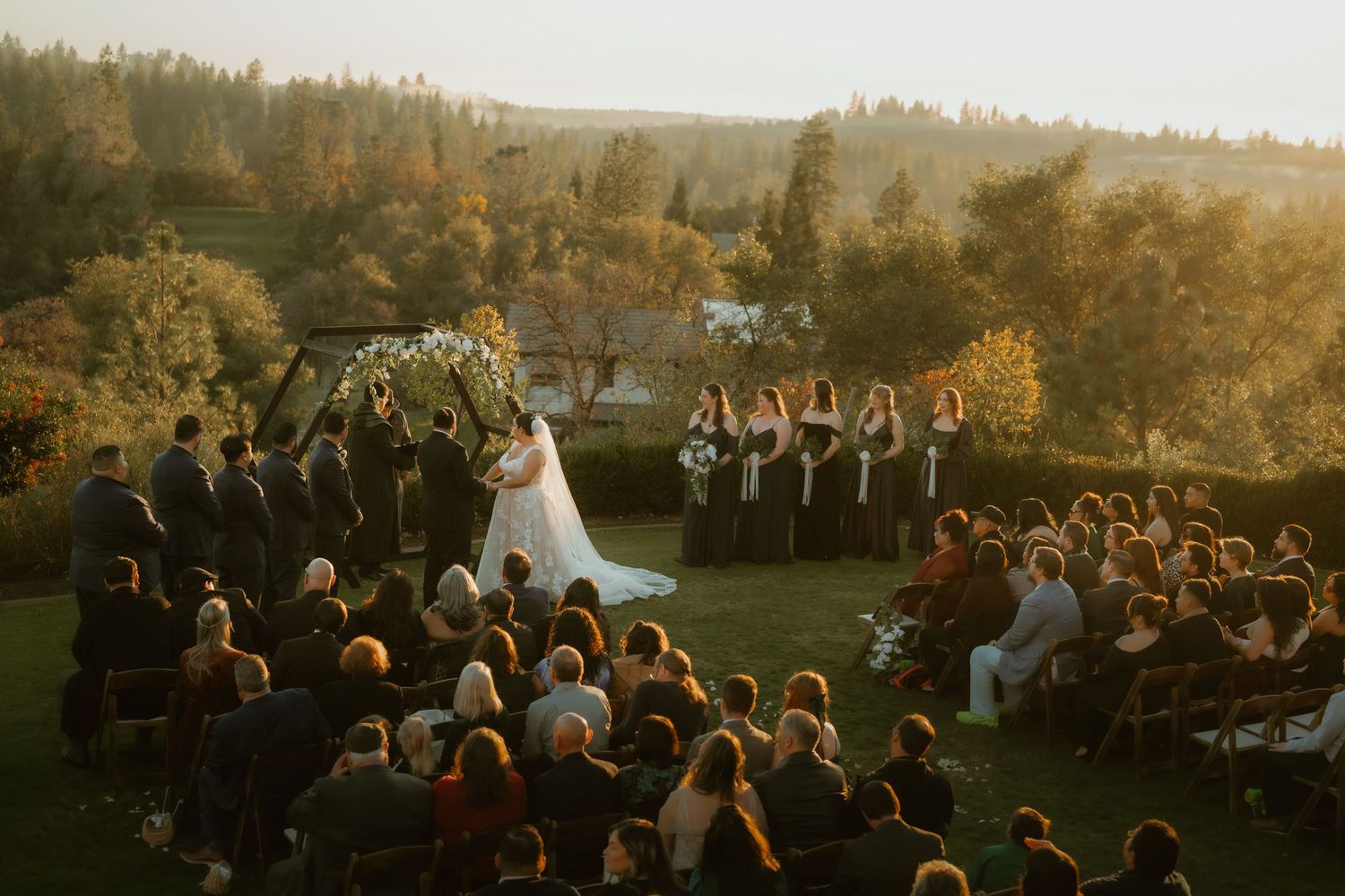 Outdoor wedding ceremony at golden hour with bridal party, guests seated on lawn, surrounded by autumn trees.