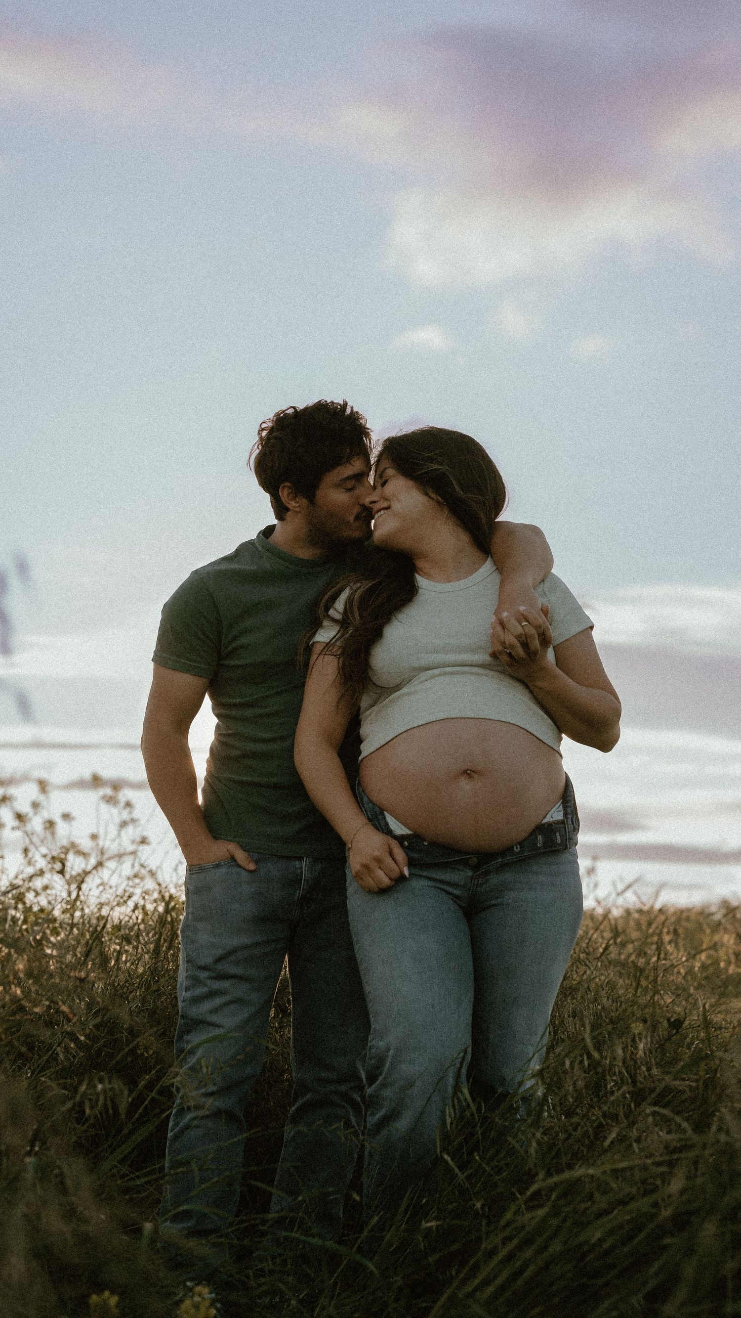 Couple embraces in field during sunset while celebrating pregnancy.