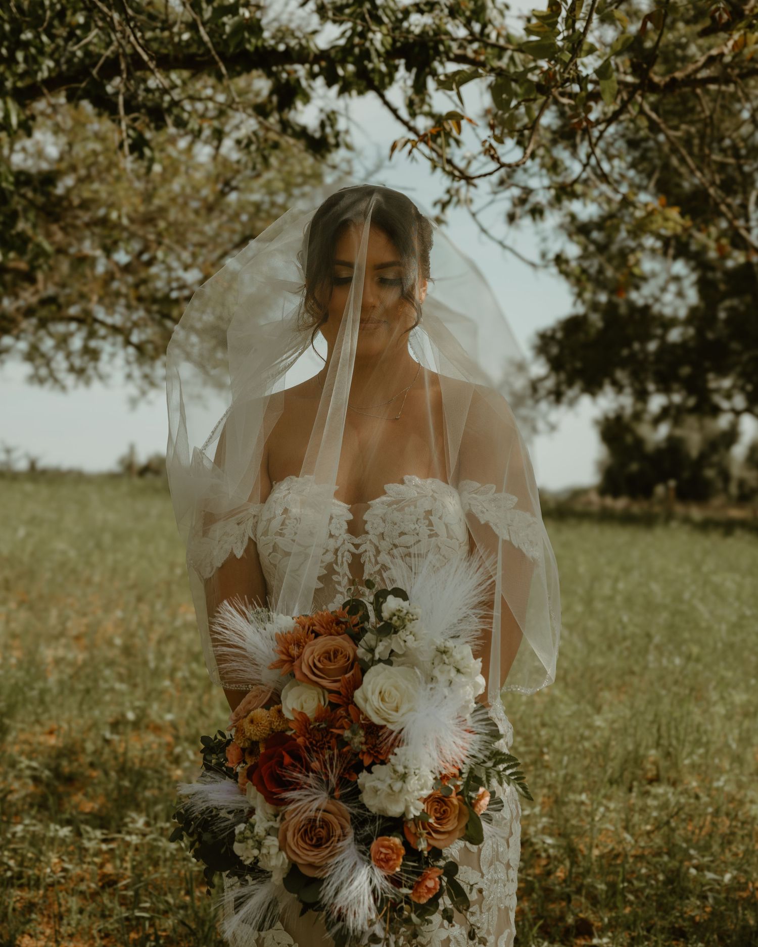 Atmospheric portrait of a bride with veil holding autumn-toned bouquet in a field.