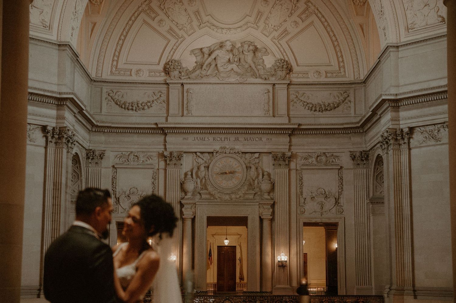 A couple shares their first dance in an ornate ballroom with classical architectural details and vaulted ceilings.