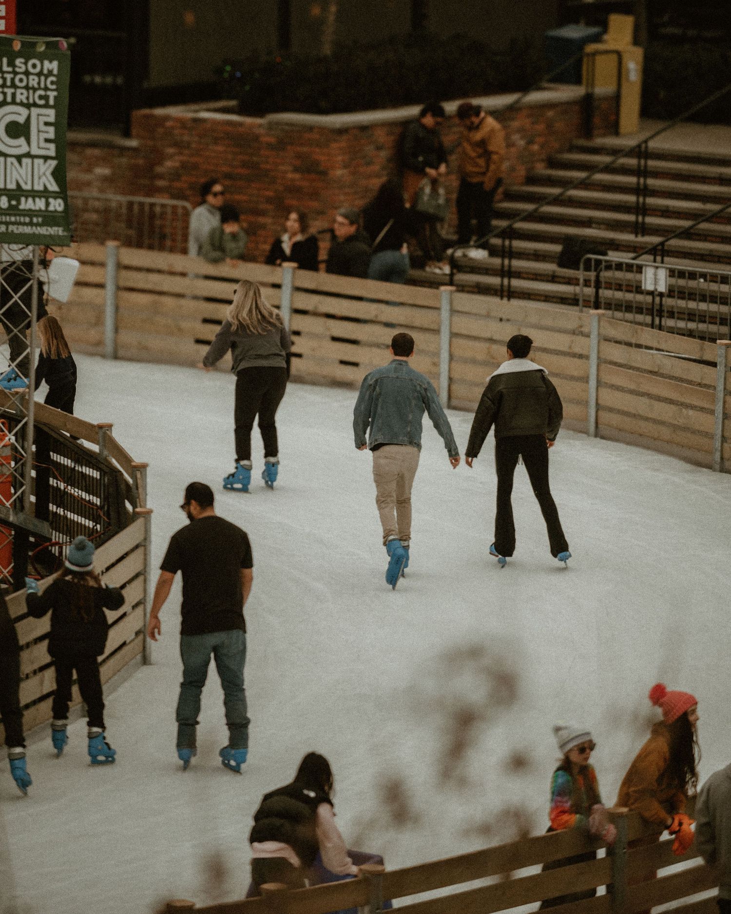People enjoying a winter ice skating rink with wooden benches and brick walls in the background.