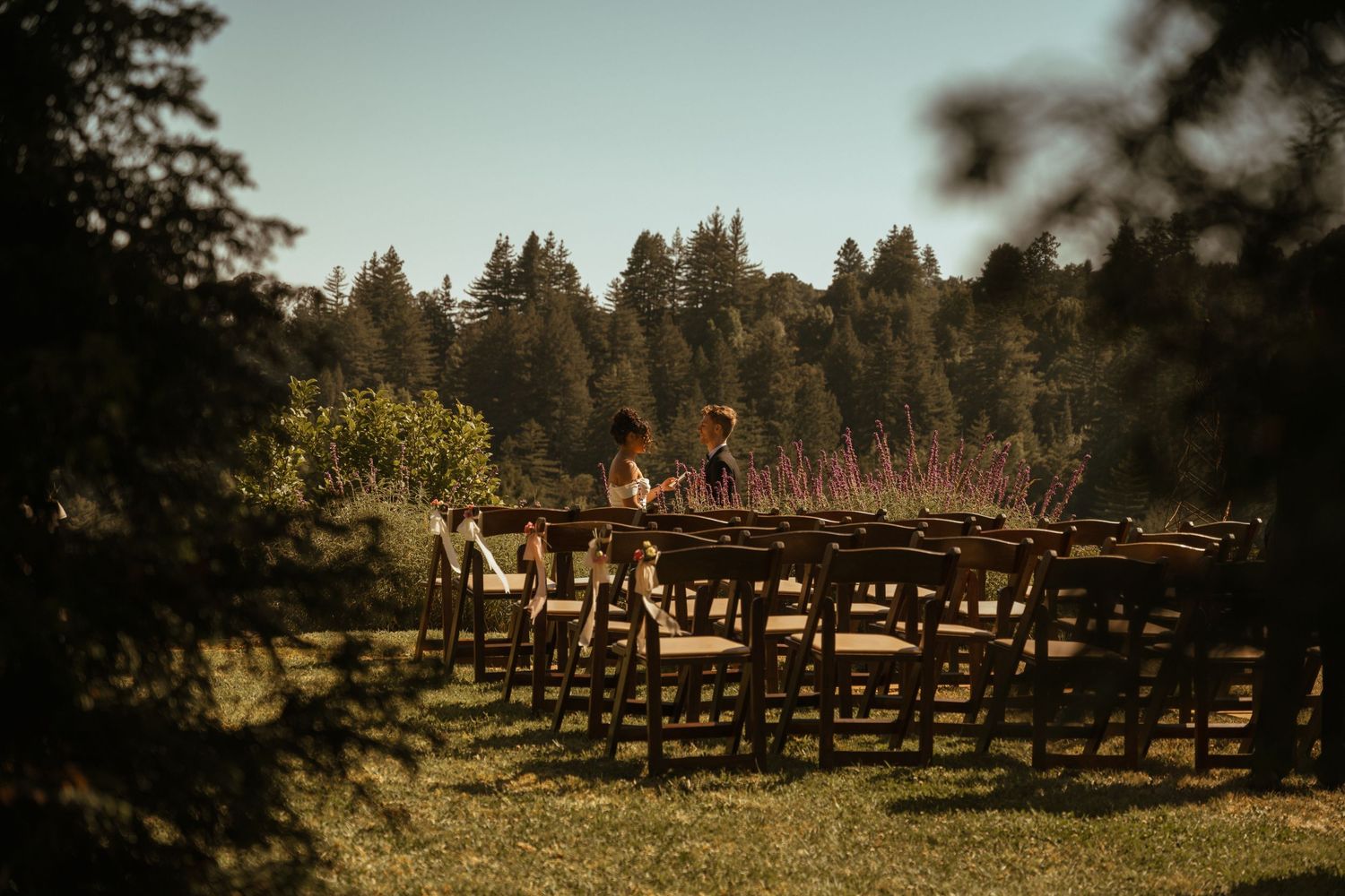 Rustic outdoor wedding ceremony setup with wooden chairs facing a forest backdrop.