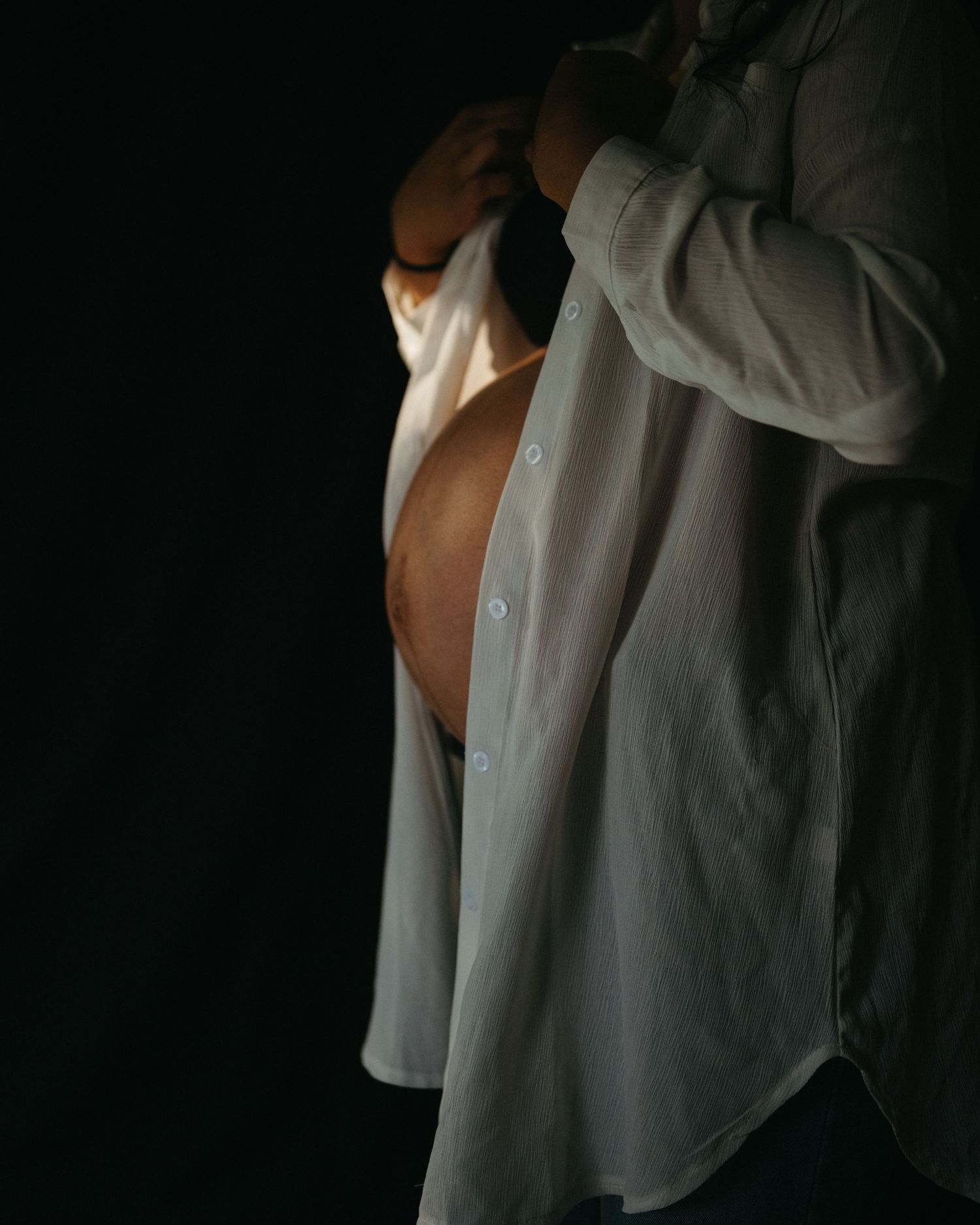 Dramatic side-lit maternity portrait with white button-down shirt against black background.