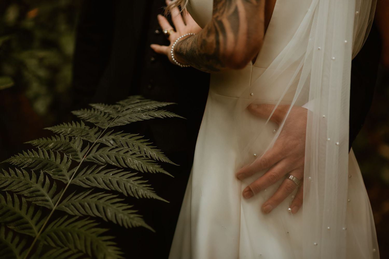 A couple's hands embrace tenderly against a backdrop of delicate ferns and white fabric in a romantic wedding photo.