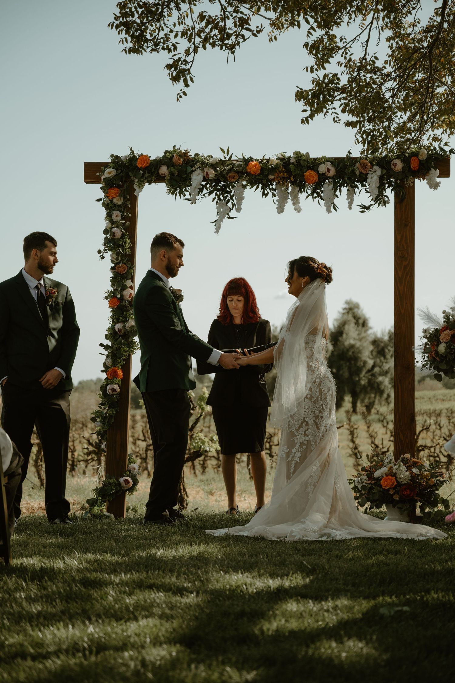 Wedding ceremony taking place outdoors under trees with guests gathered in formal attire.