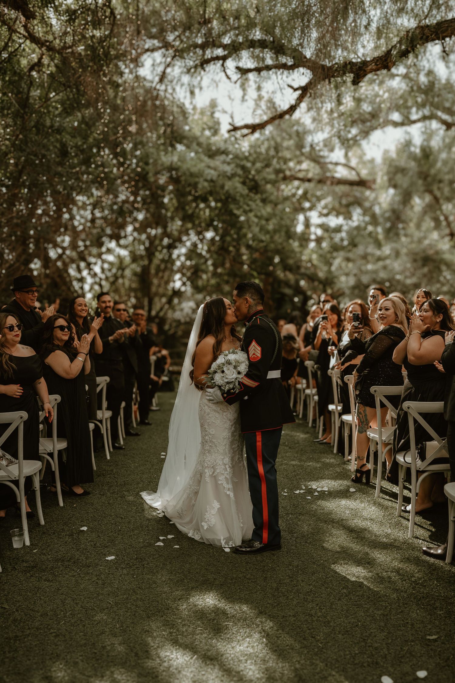Wedding ceremony guests seated outdoors under trees with a bride and Marine groom at the altar.