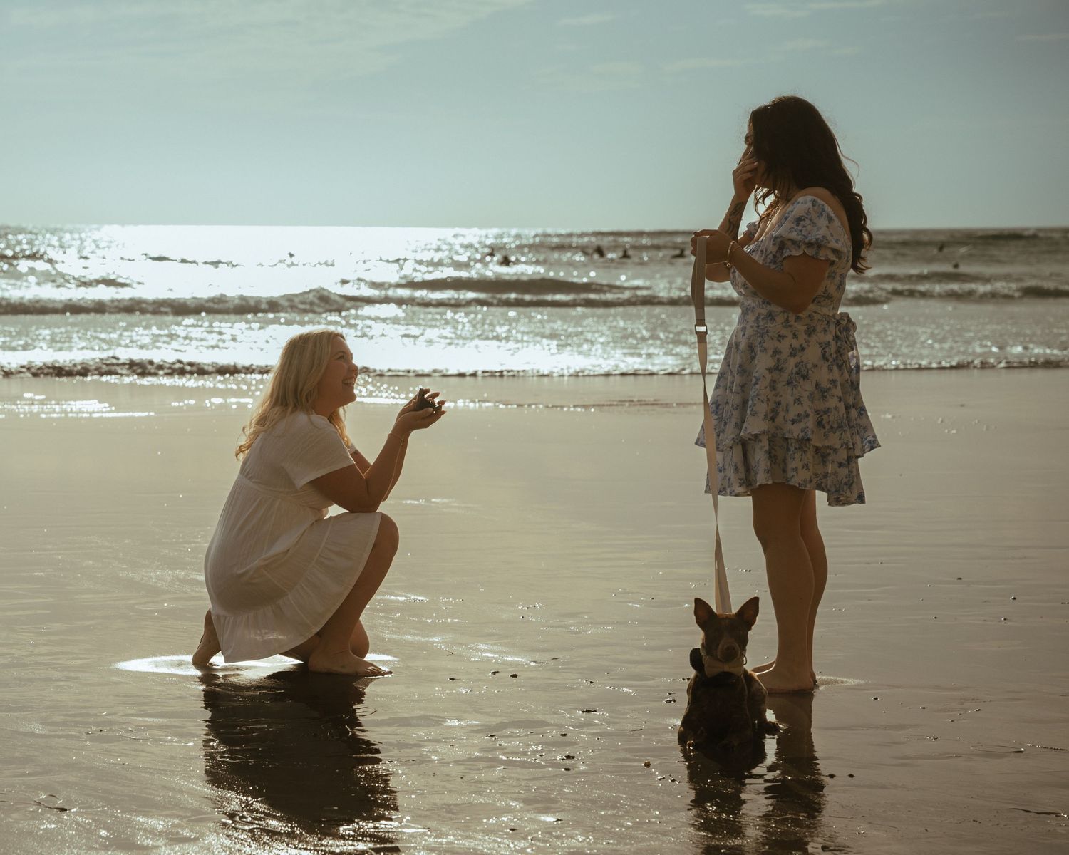Two women and a dog on a sunny beach, one kneeling as waves lap the shore.