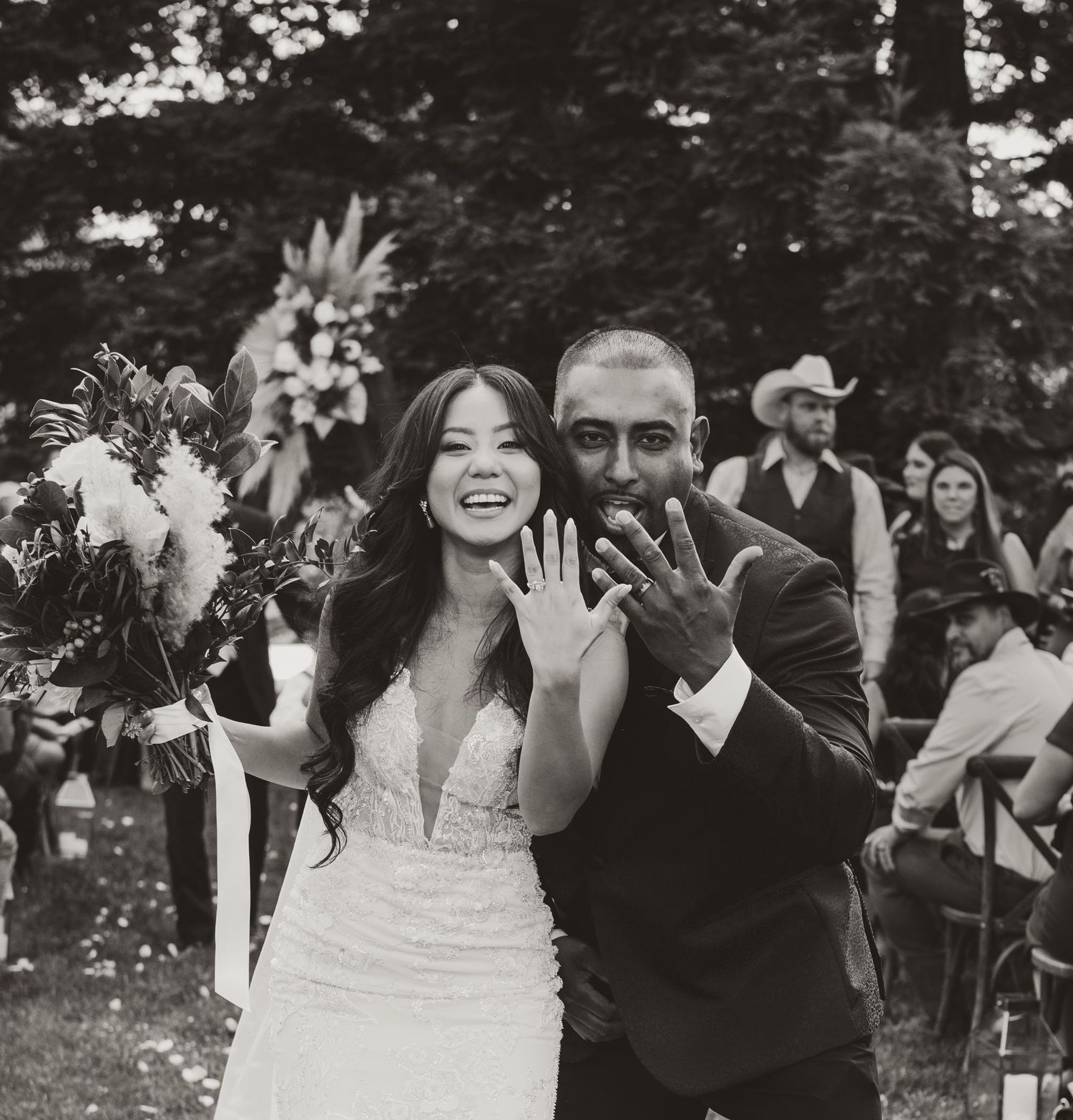 Newlyweds showing off their wedding rings with joyful expressions during an outdoor garden ceremony.