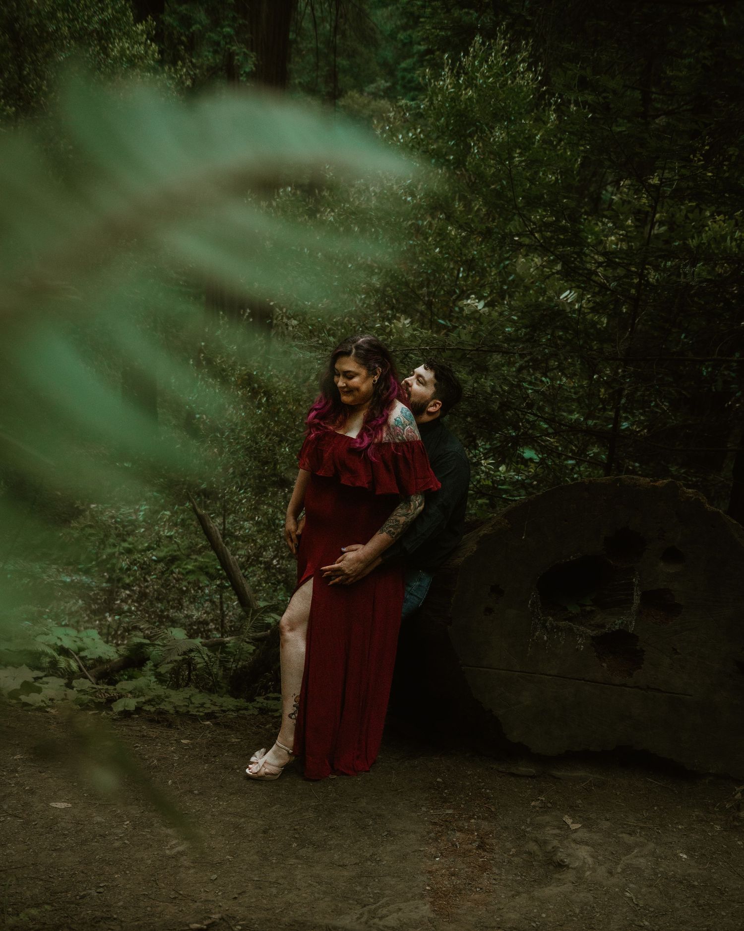 Couple in a romantic forest setting with one wearing a flowing burgundy dress amid lush greenery.