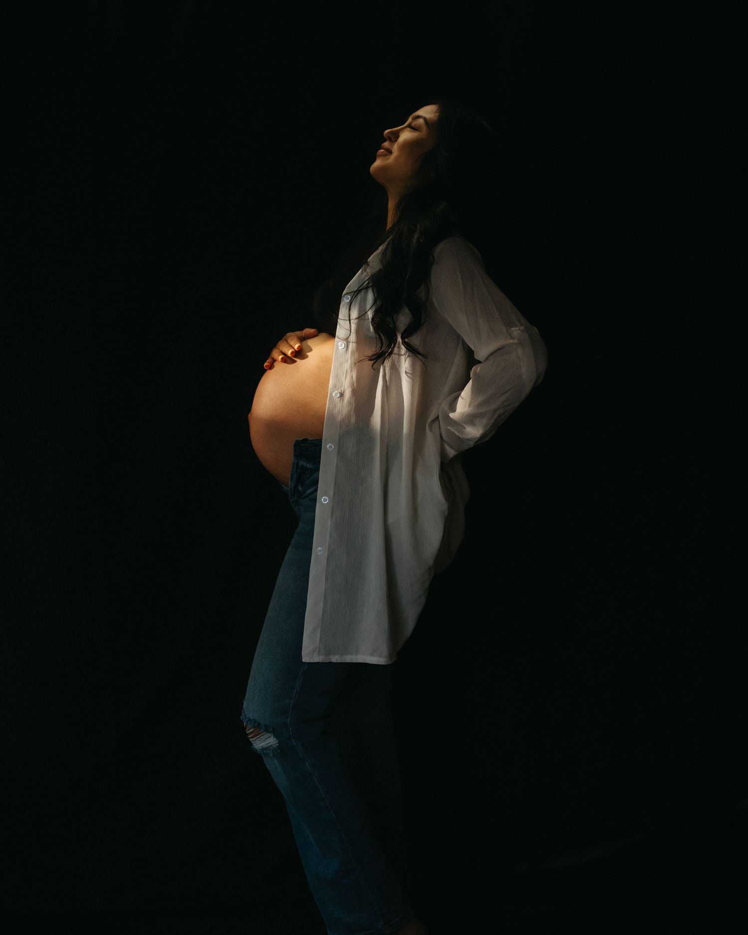 Artistic silhouette maternity portrait against dark background with dramatic lighting.