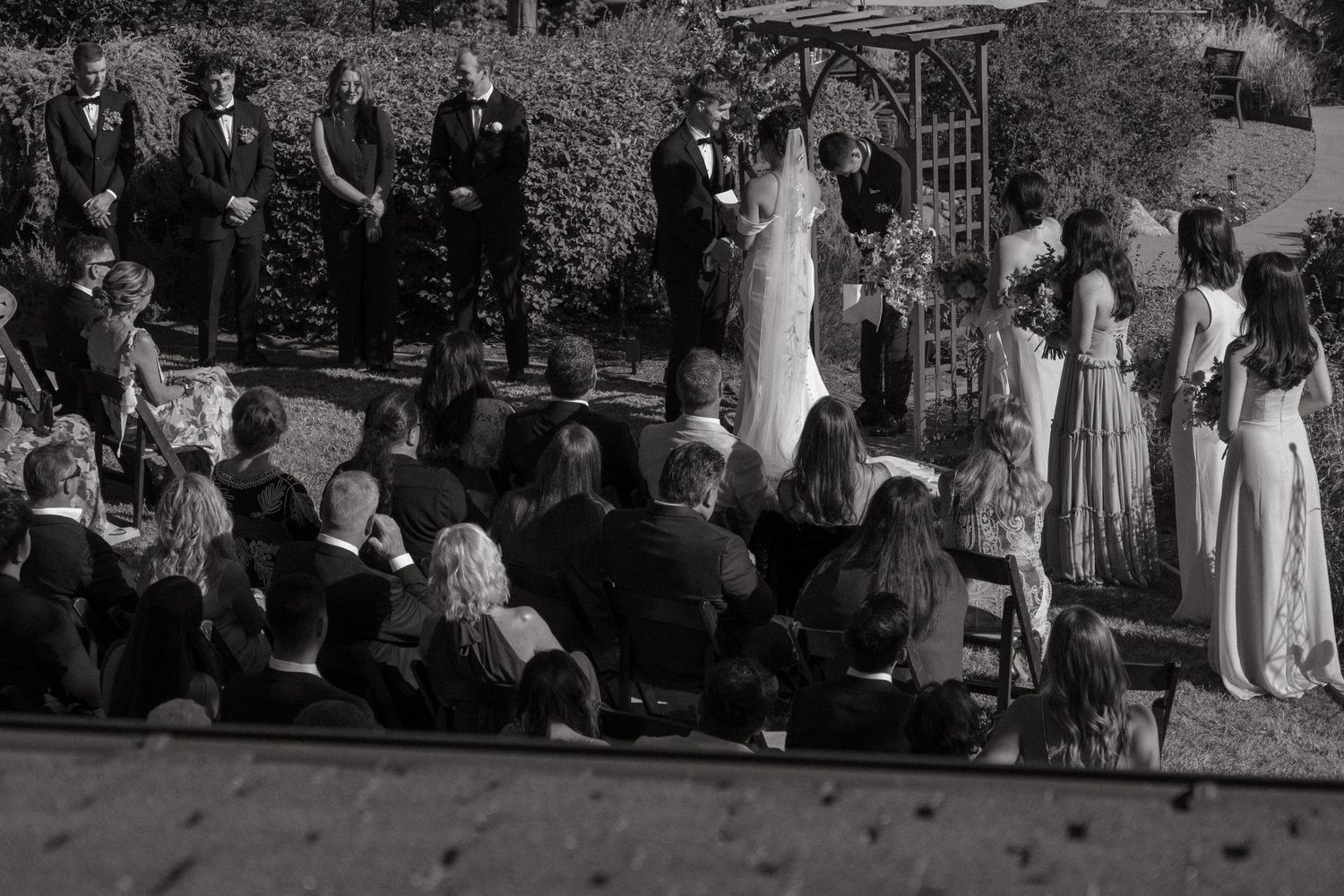 Black and white photograph of an outdoor wedding ceremony under an arbor.