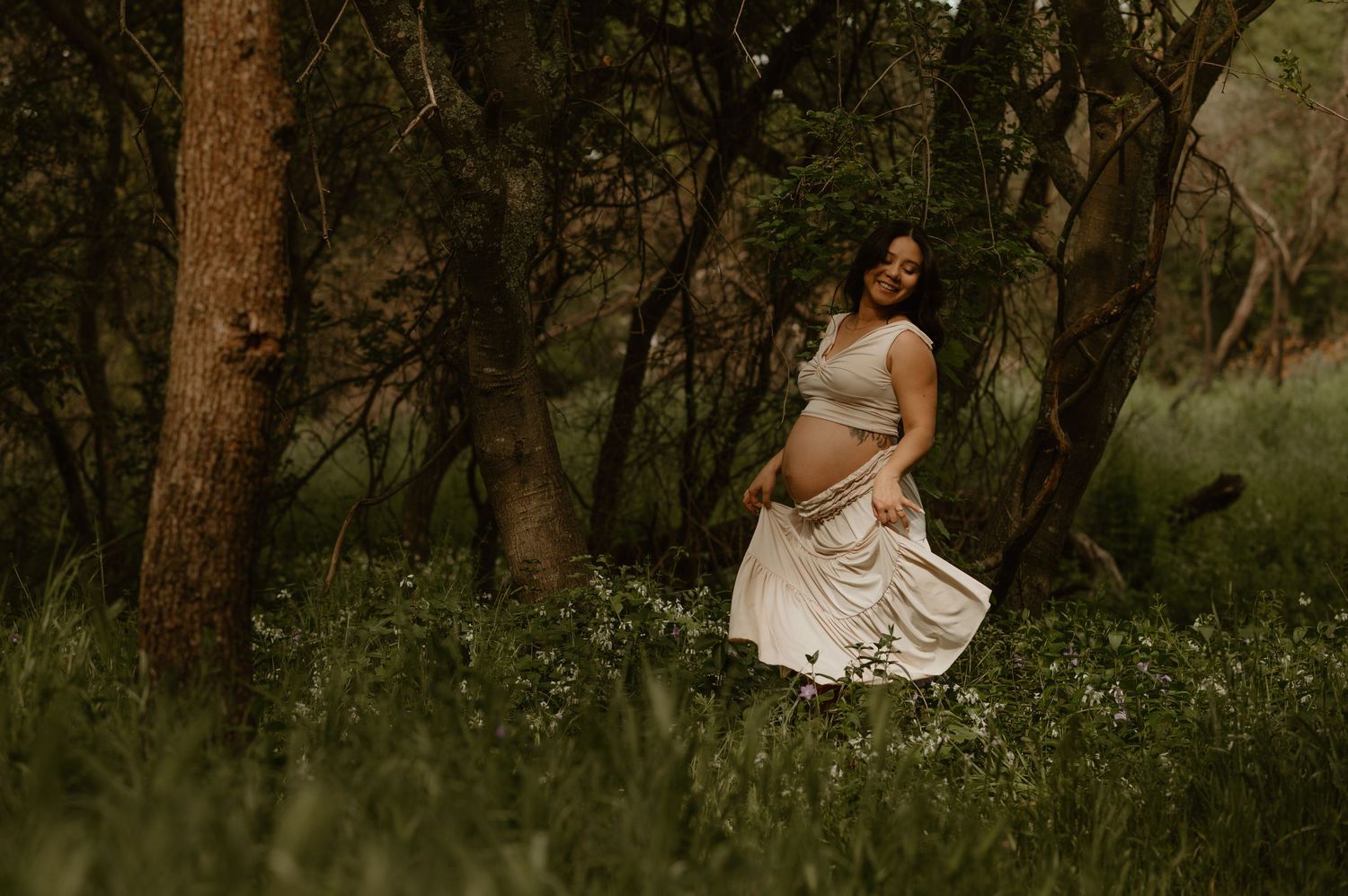 Romantic maternity photo in a wooded area with white wildflowers and natural lighting.