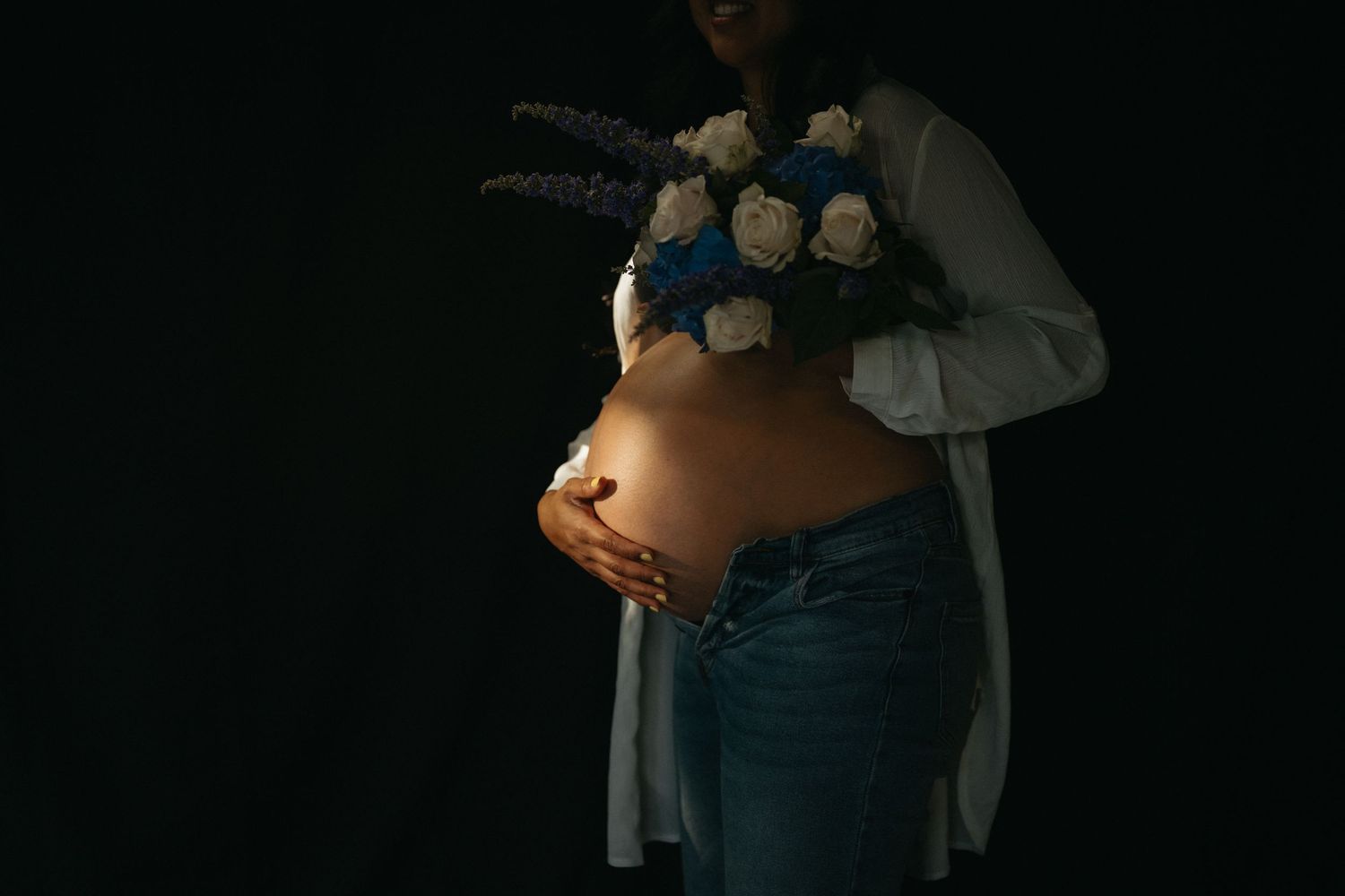 Artistic maternity photo showing a pregnant belly with white and blue flowers in low light.