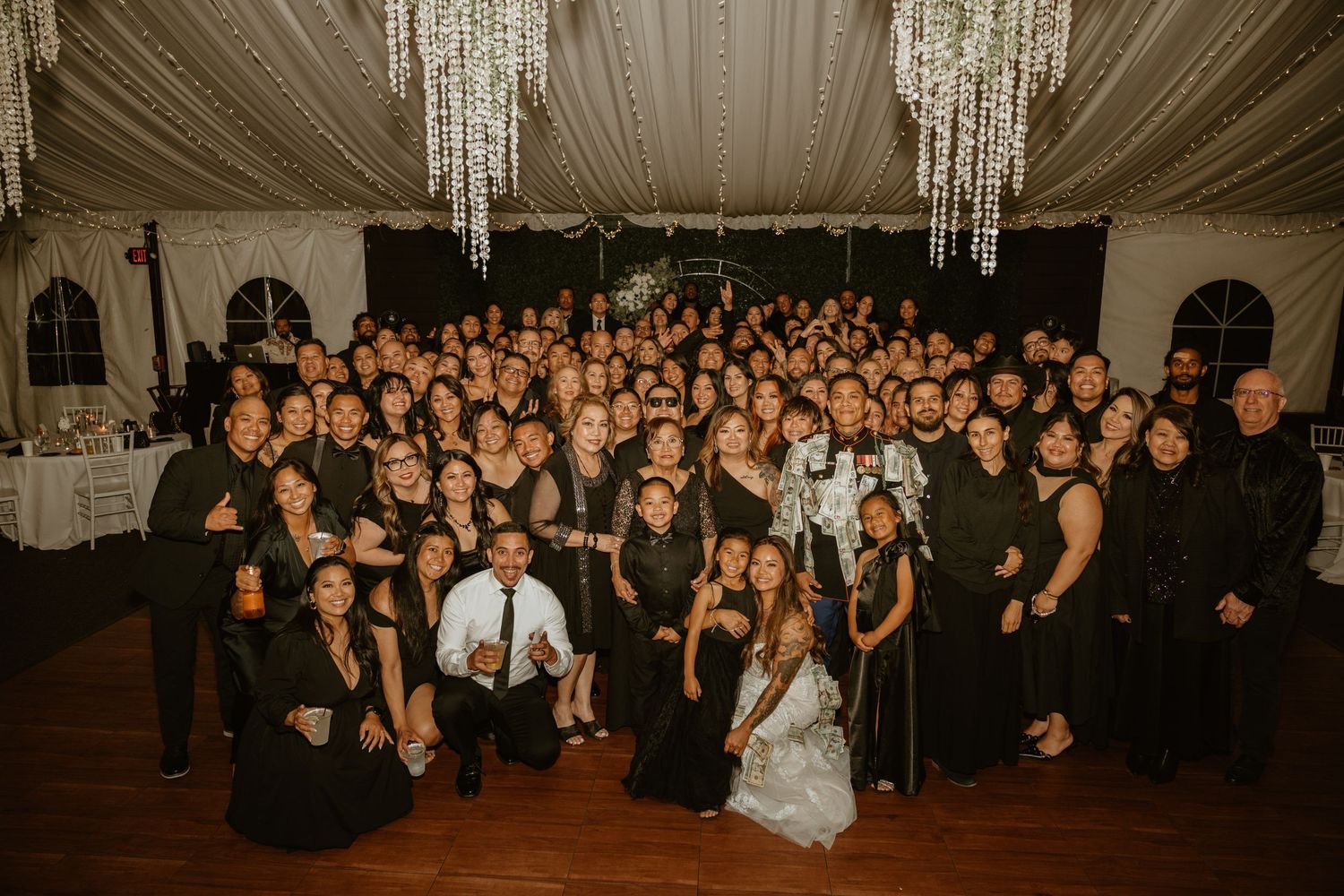 Large group photo of wedding guests in an elegant reception hall with crystal chandeliers.