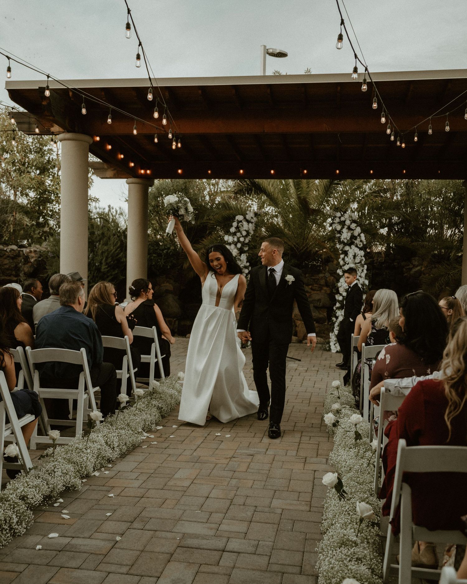 Newlyweds celebrate walking down the aisle under string lights at an outdoor ceremony.
