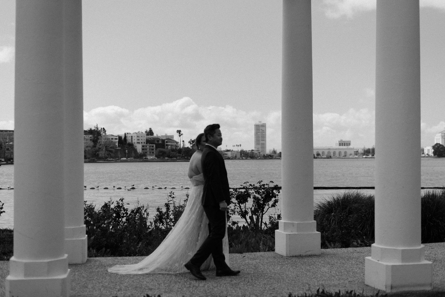 A couple walks between white columns with a scenic waterfront view.