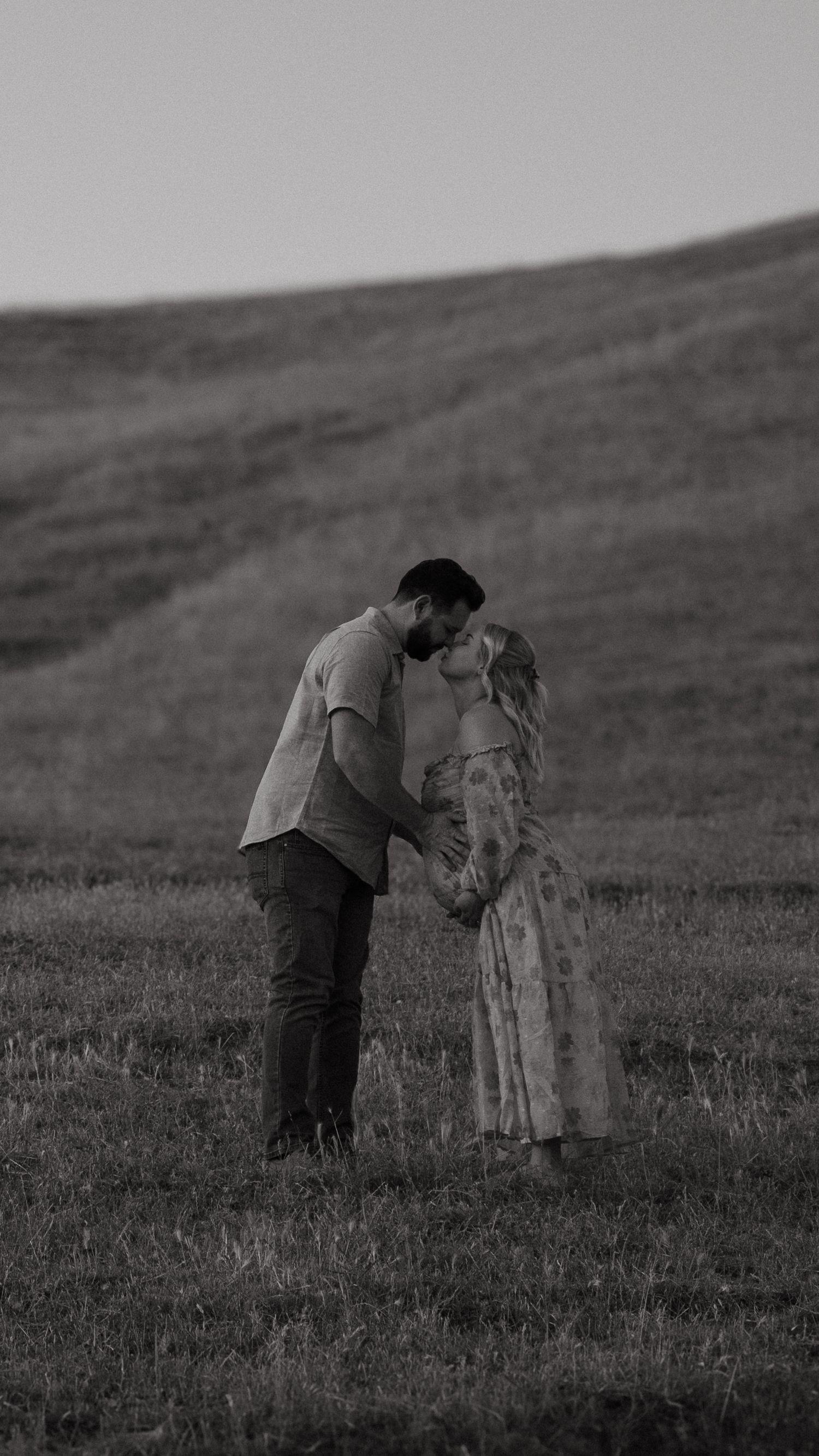 Intimate black and white portrait of couple embracing on grassy hillside.