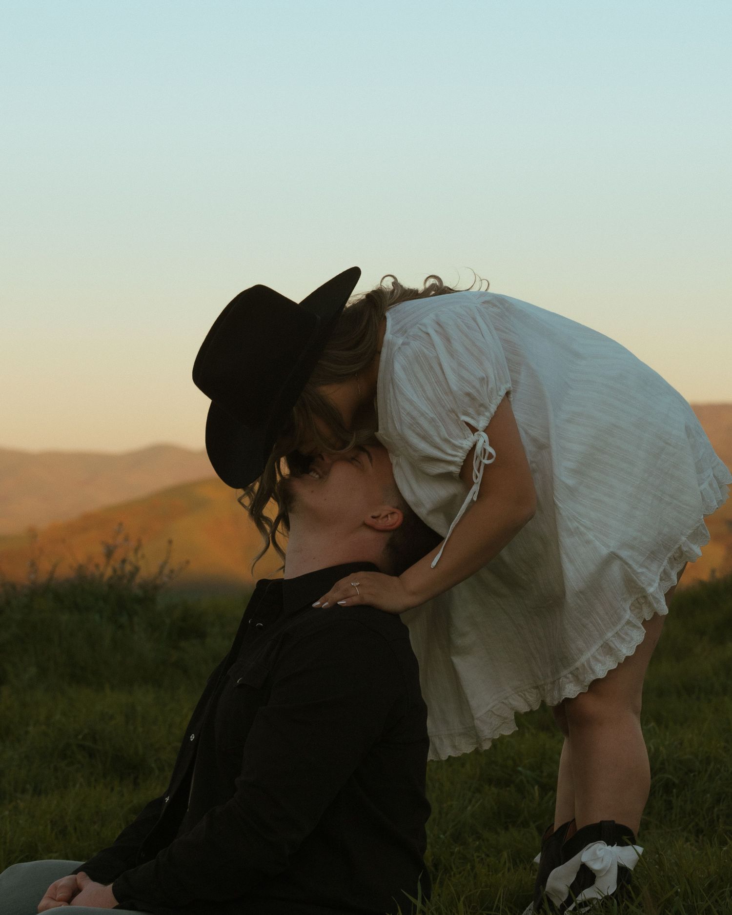 Couple sharing a romantic kiss at golden hour on a grassy hill, woman in white dress and man in black cowboy hat.
