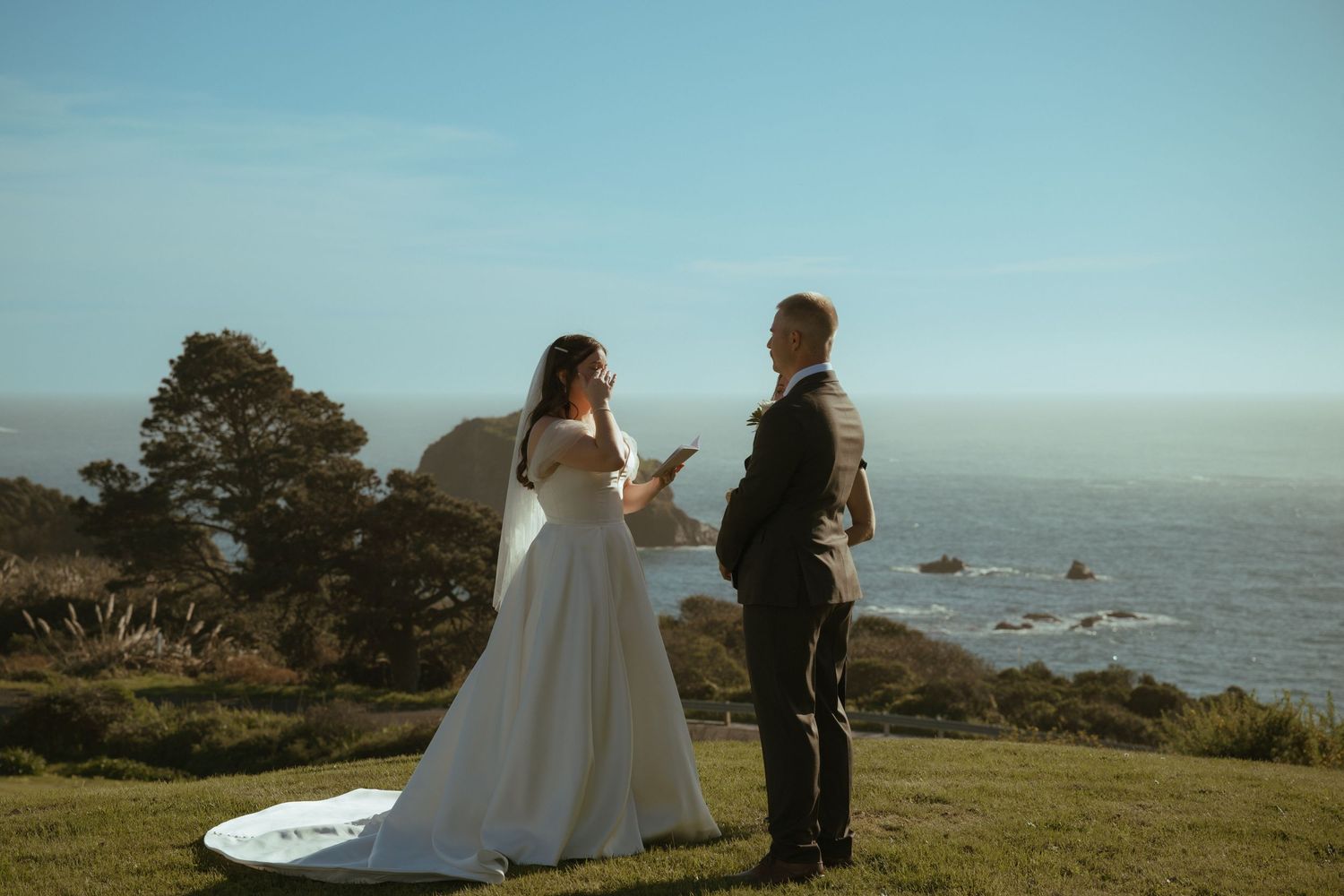 Bride and groom exchange vows outdoors on a cliffside overlooking the ocean on a sunny wedding day.