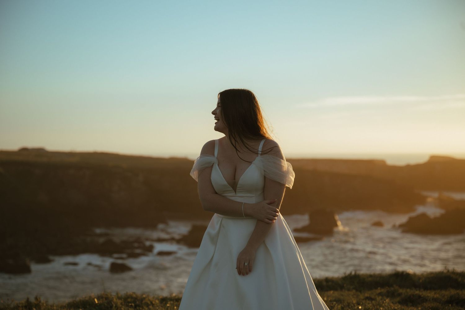Bride in white off-shoulder gown standing on coastal cliffs at golden hour sunset.