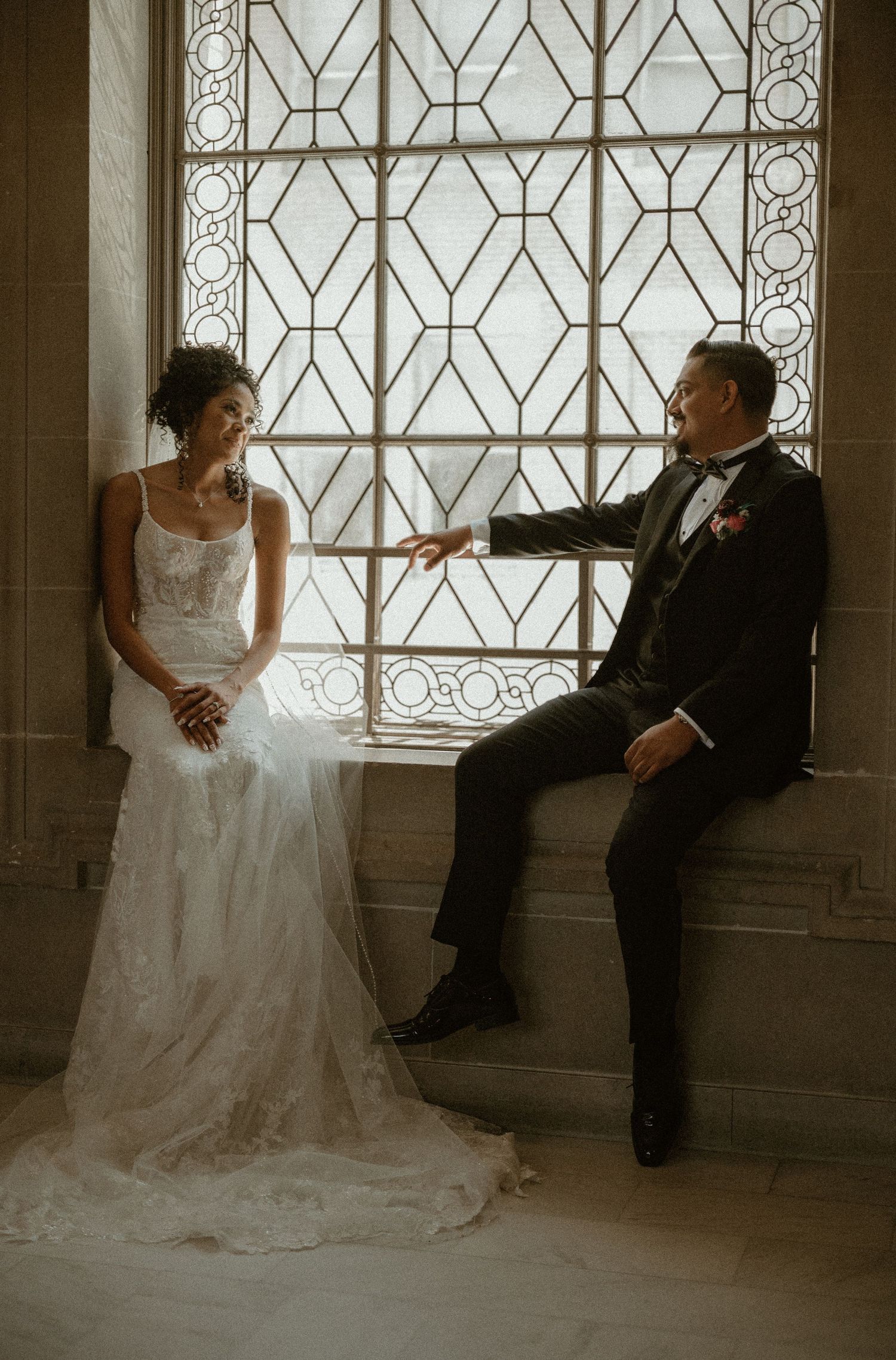 A couple poses by an elegant geometric window in formal wedding attire inside a historic building.