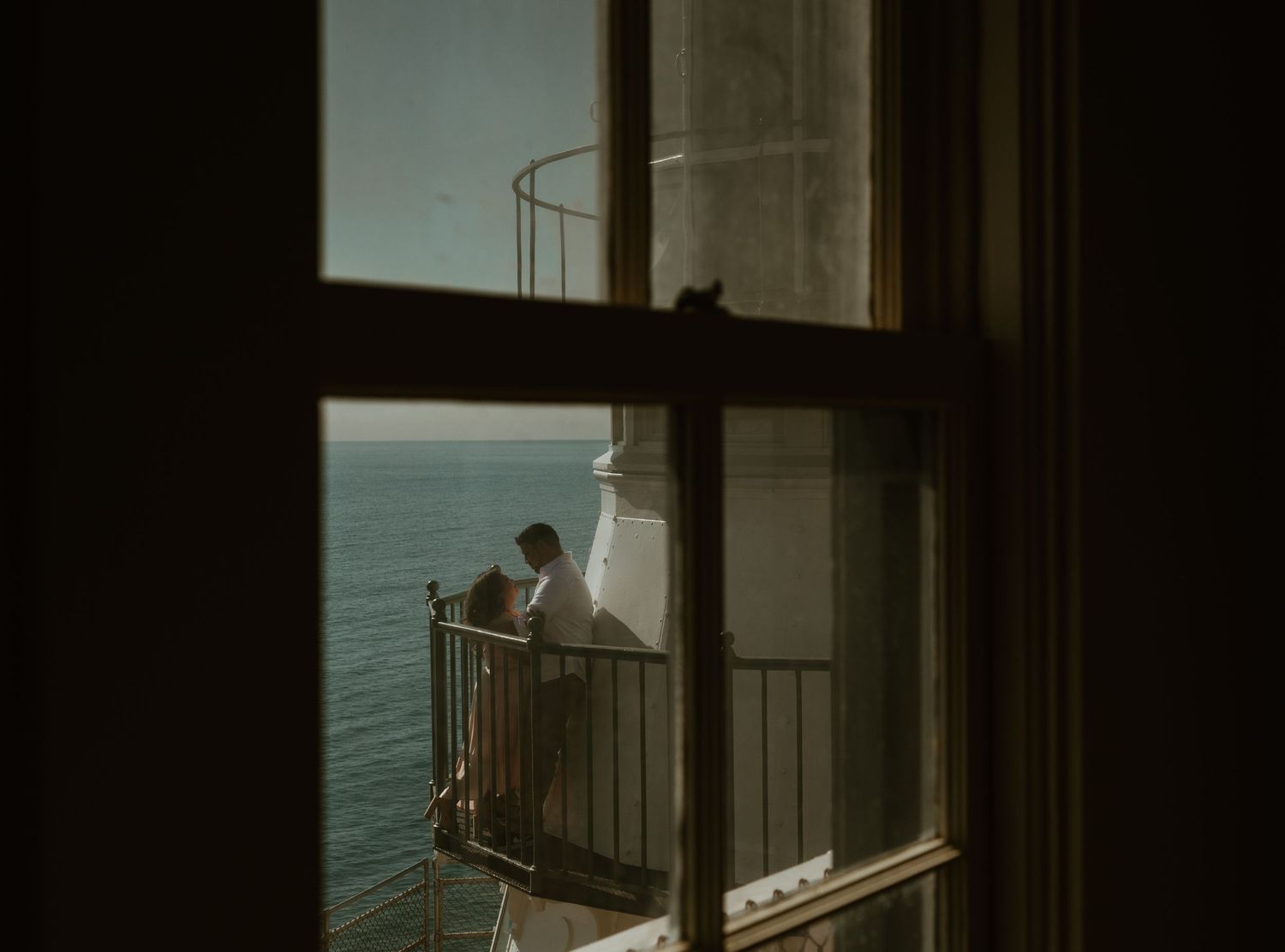 A person stands at a balcony railing overlooking a calm body of water during sunset or sunrise.
