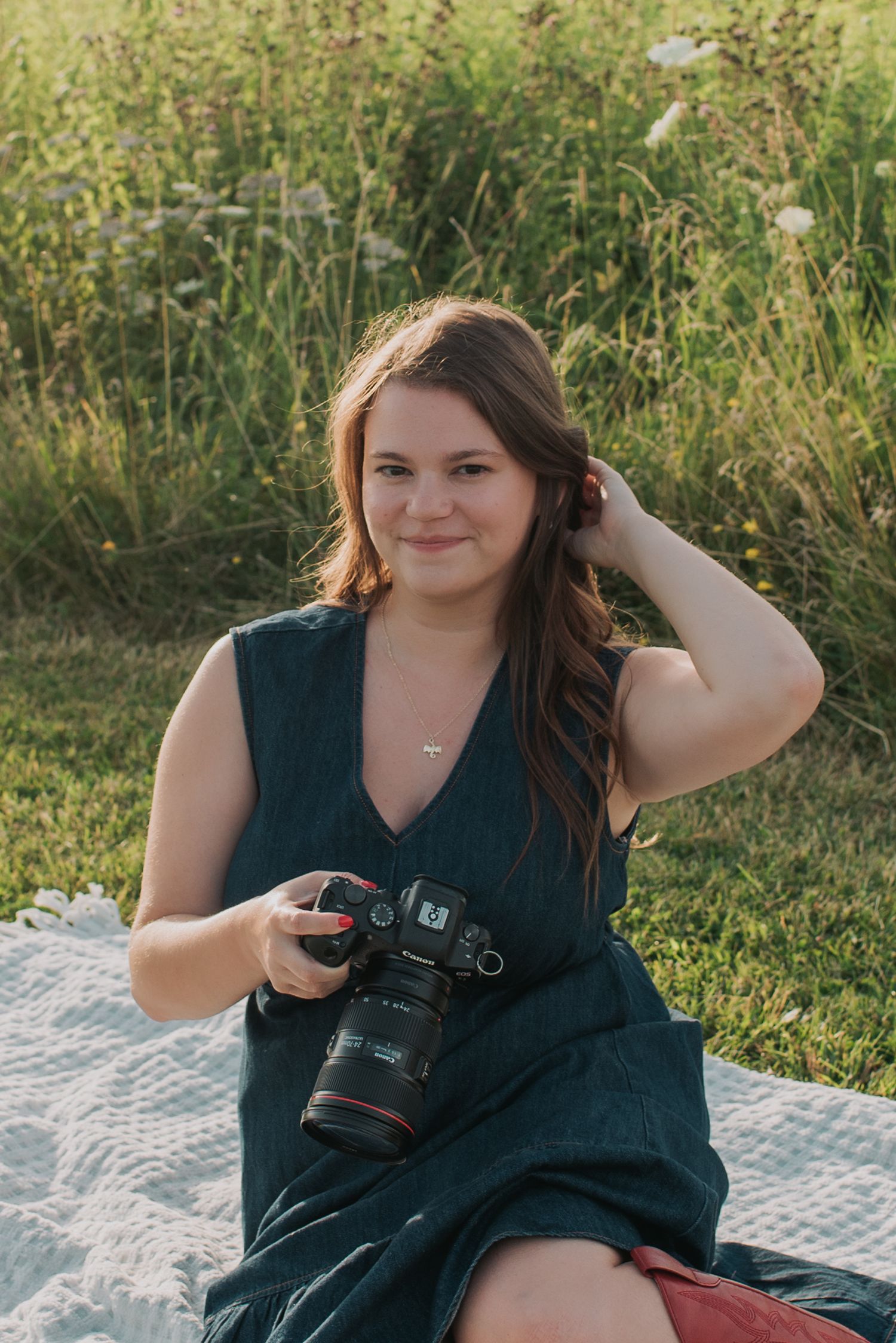 Smiling Altoona photographer in navy dress poses with camera on picnic blanket in summer field.