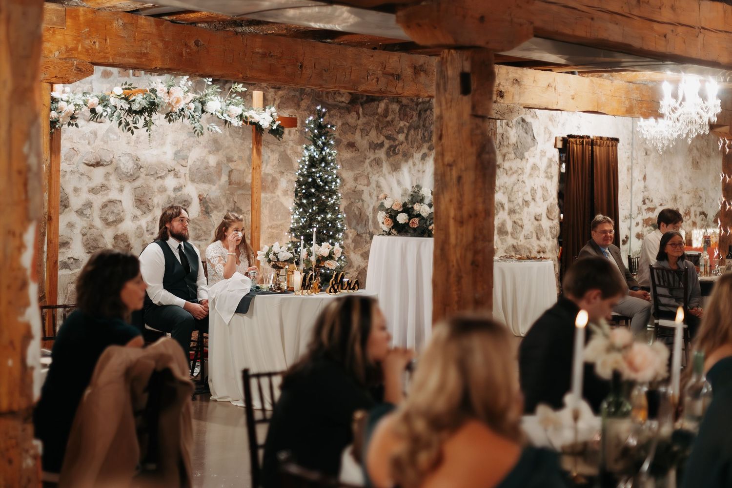 Wedding party celebrates in a rustic brick-walled reception venue decorated with twinkling lights and floral arrangements.