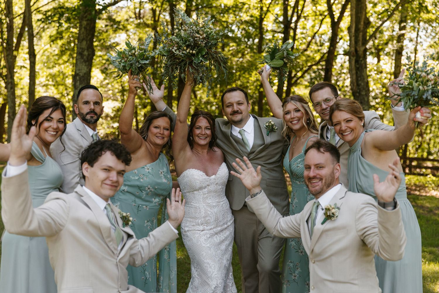 Joyful wedding party celebrating outdoors in forest, bride and groom surrounded by bridesmaids in sage green and groomsmen in tan suits.
