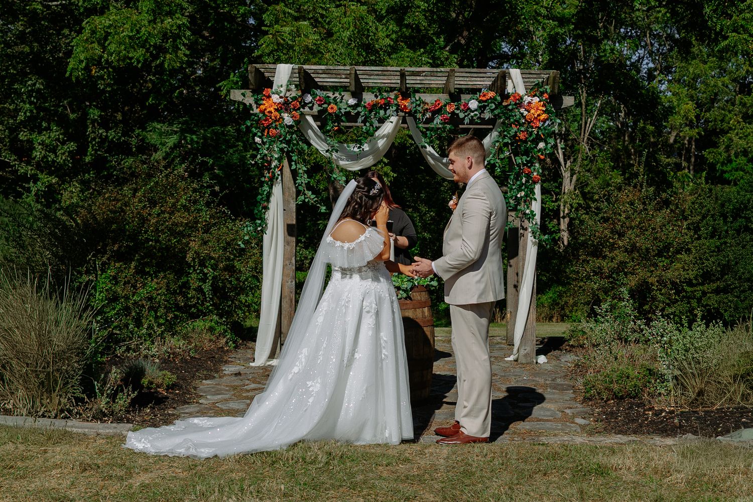 A wedding ceremony takes place under a rustic wooden arch decorated with flowers and greenery in an outdoor garden setting.