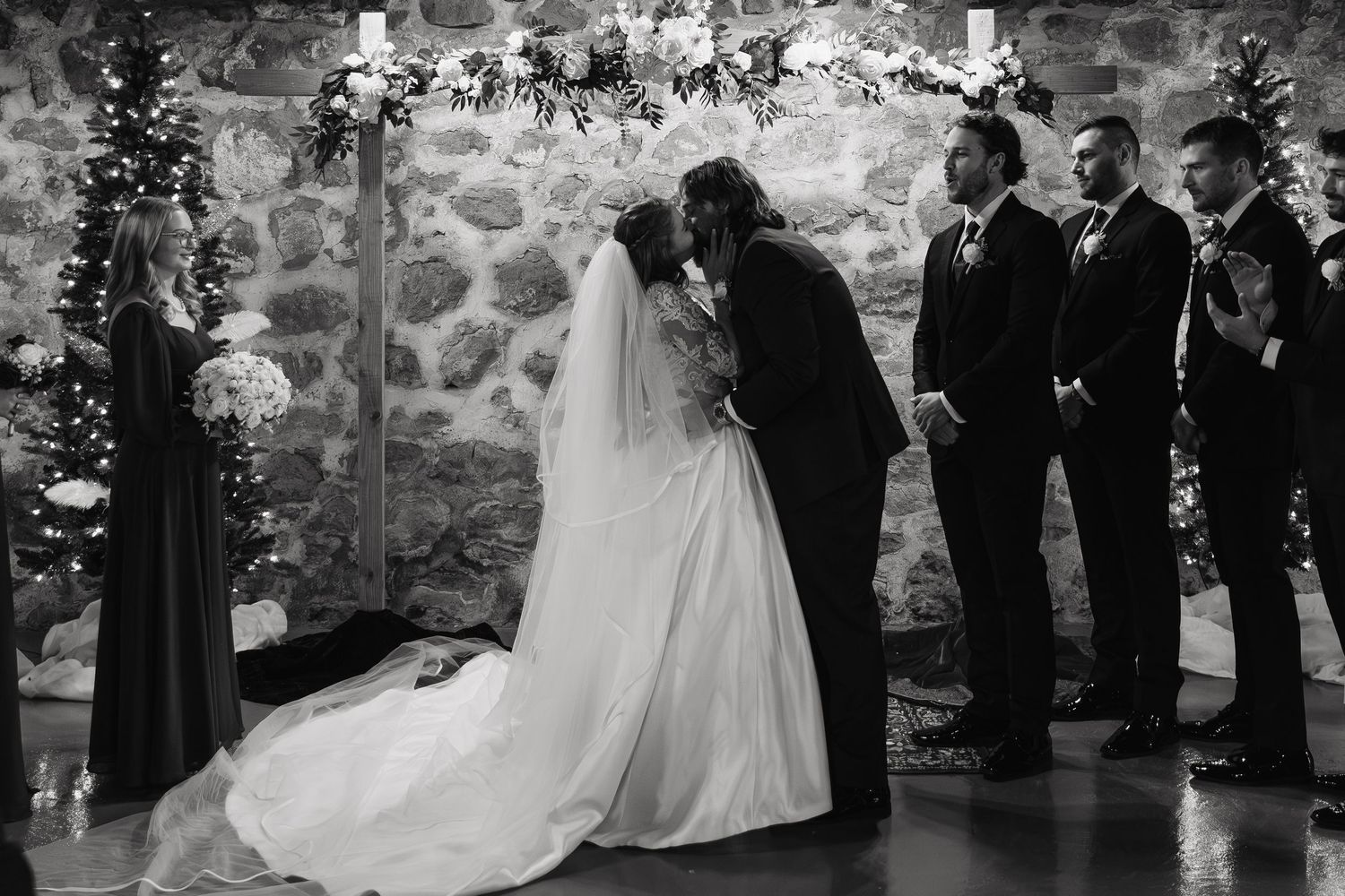 A black and white wedding ceremony moment captured against a rustic stone wall with the wedding party standing nearby.