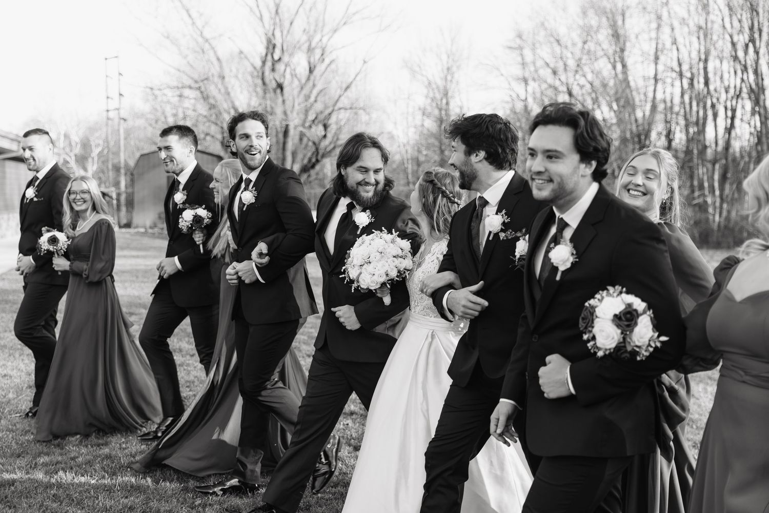 A joyful wedding party walks together outdoors in black and white, carrying bouquets and laughing.