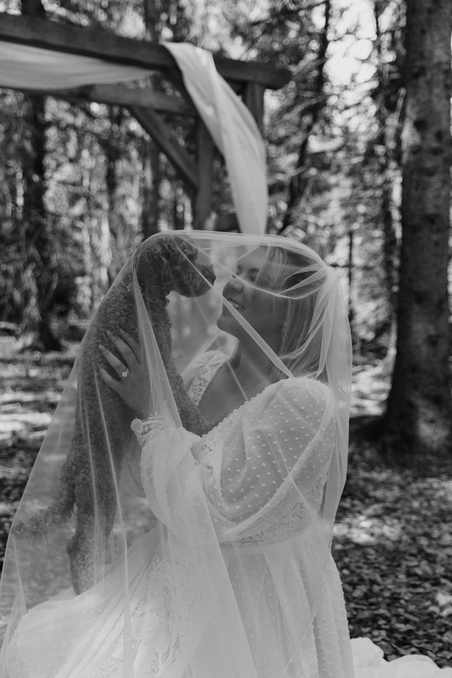 Black and white photo of bride and groom sharing a kiss under a veil in a forest wedding ceremony.