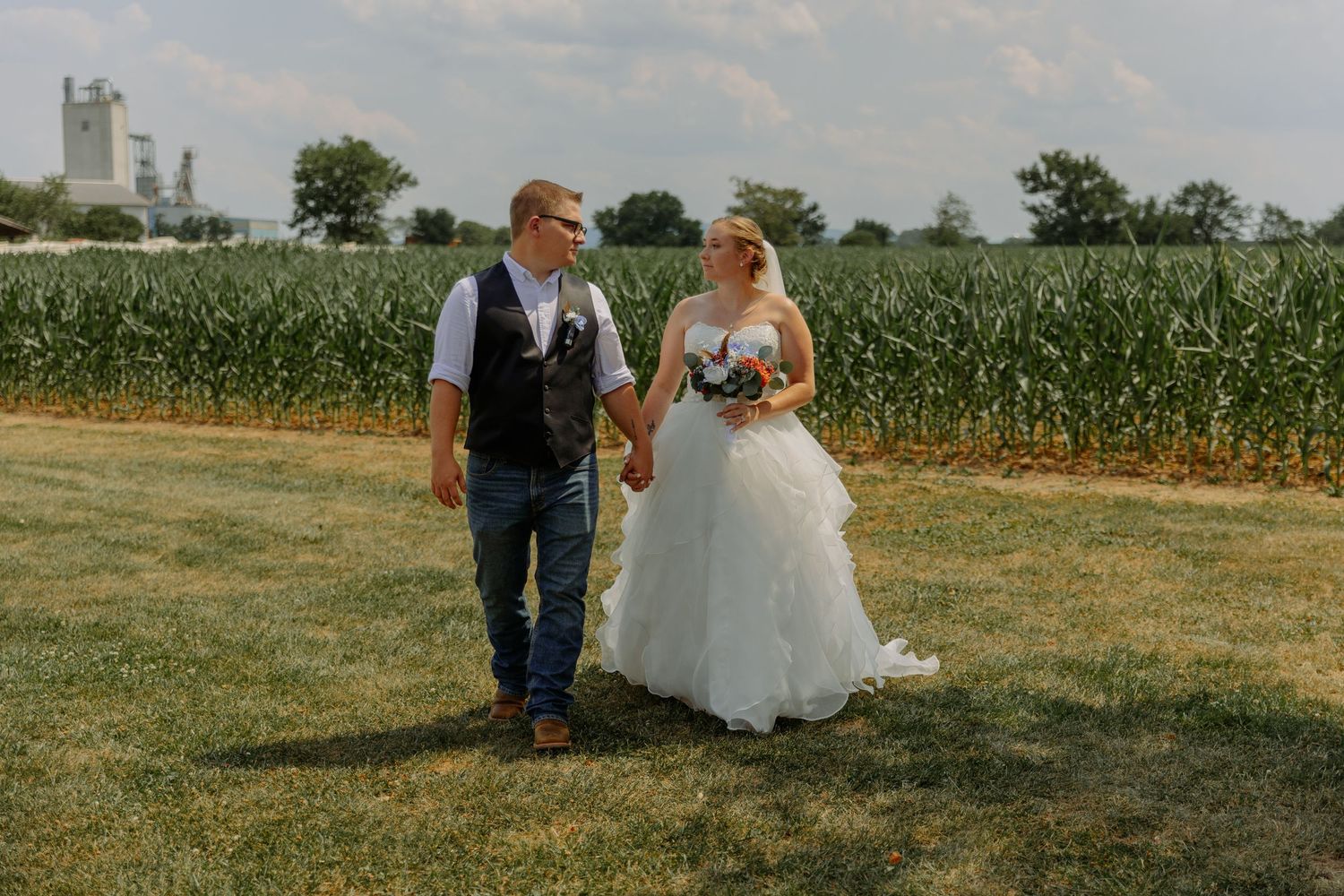 Couple walks hand in hand through rural cornfield setting in wedding attire.